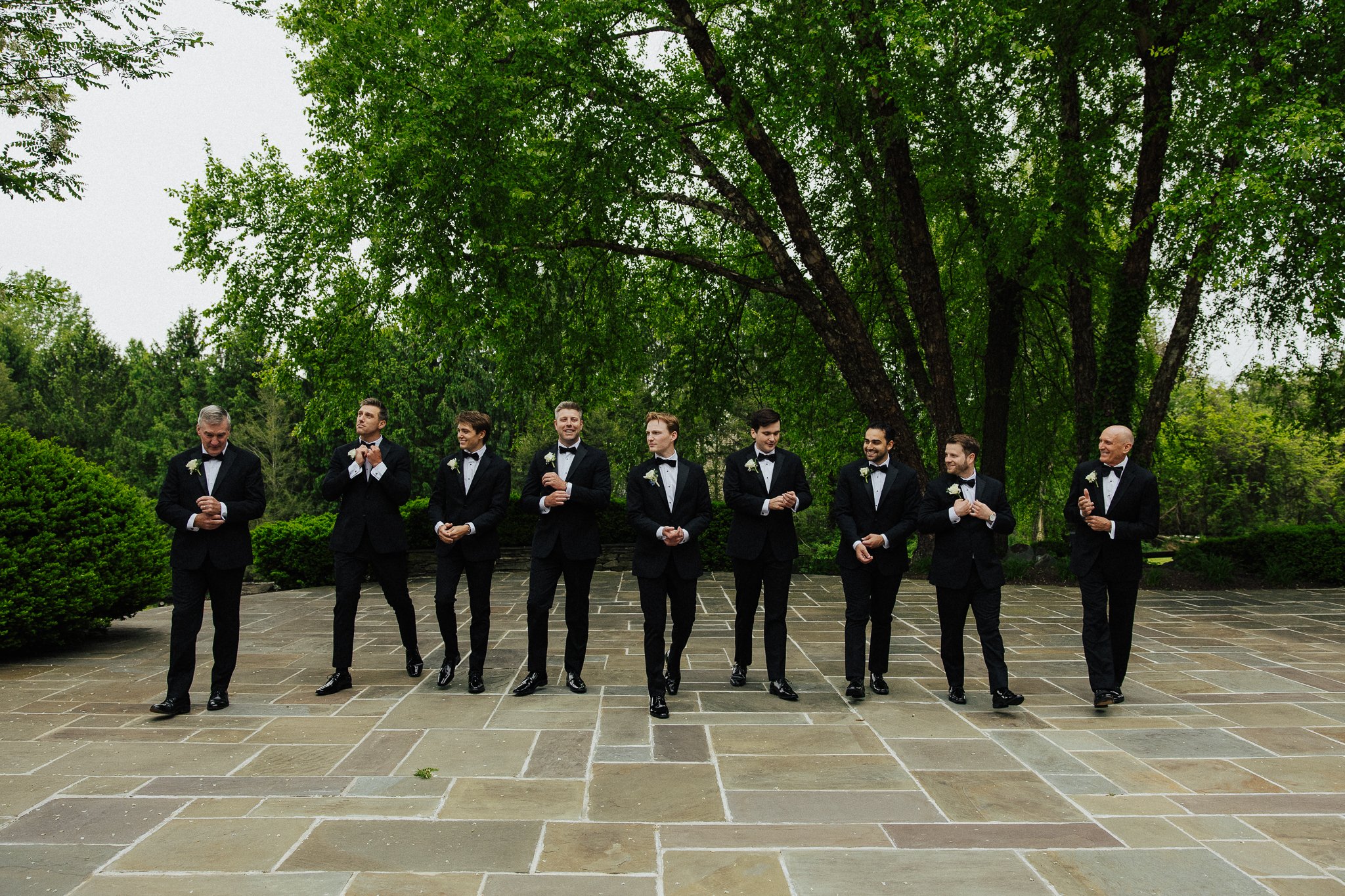 A group of nine men dressed in black tuxedos and white shirts standing in a line outdoors under a large leafy tree. They are smiling and clapping, possibly at a formal event or wedding.