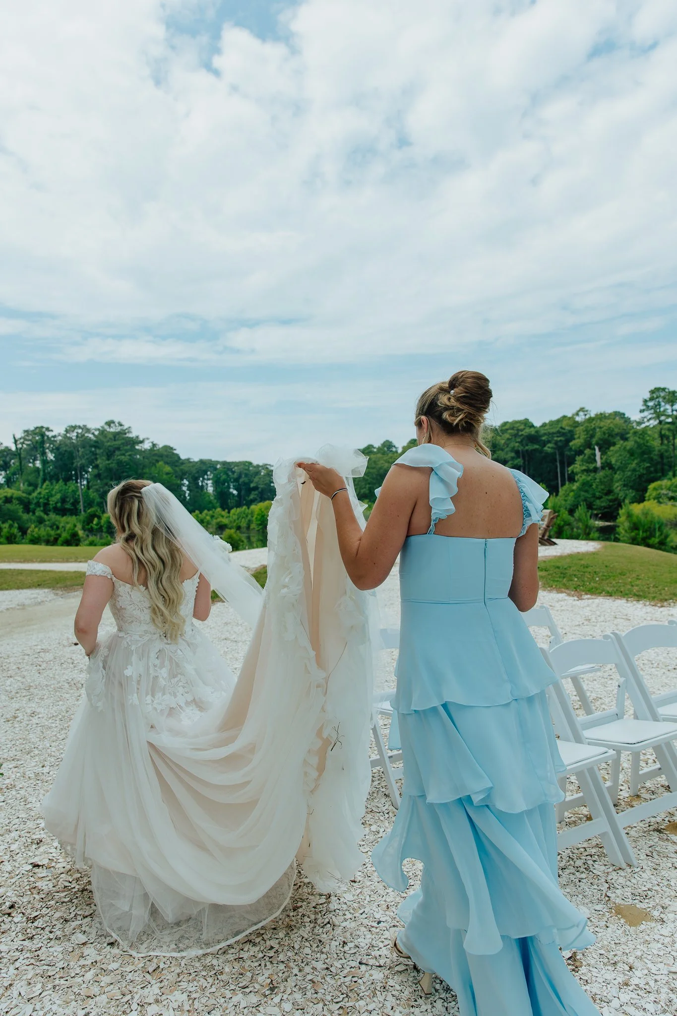 Women in dresses, likely at a wedding, outdoors with chairs and trees under a cloudy sky.