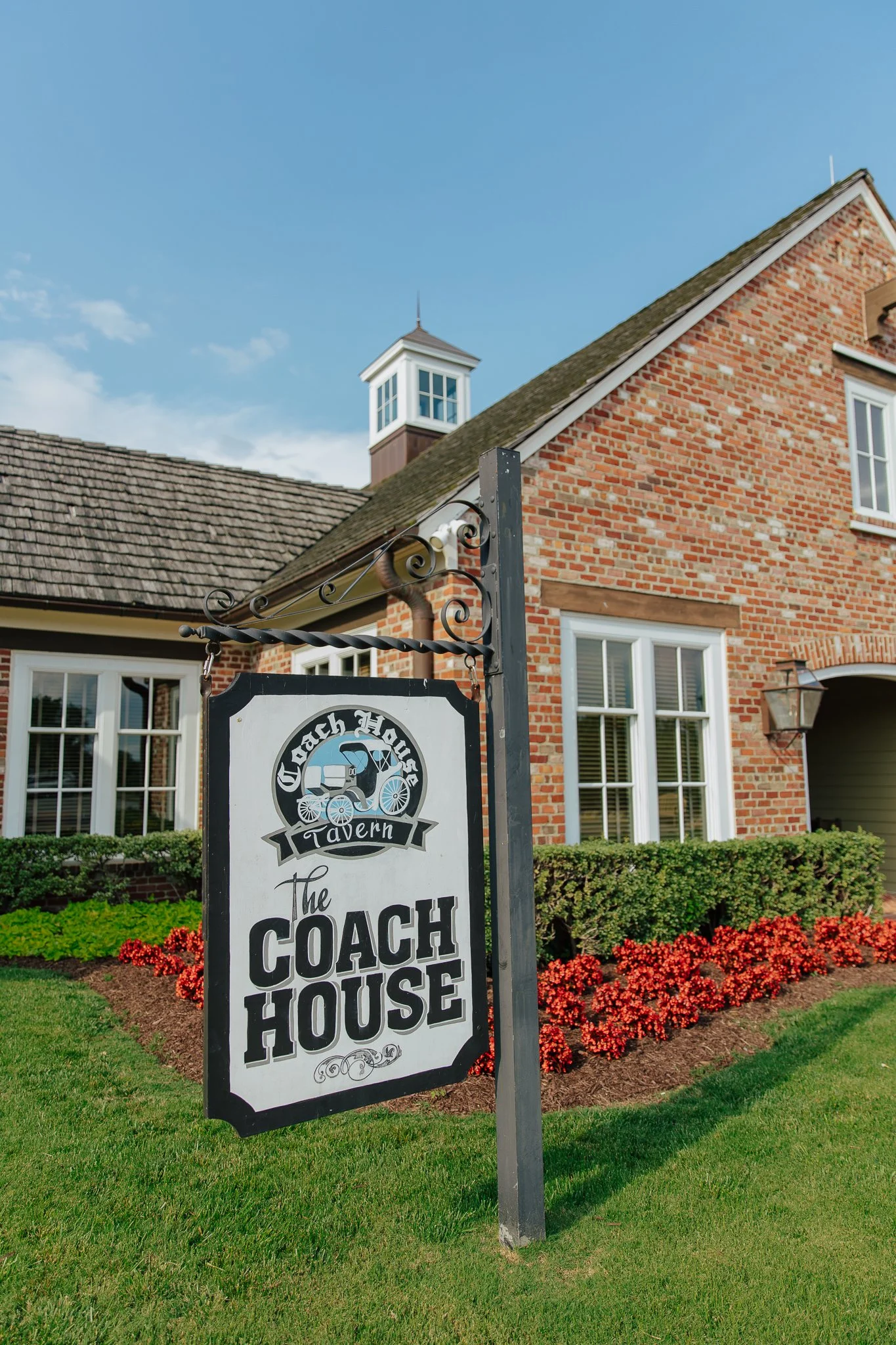 A sign for The Coach House tavern in front of a red brick building with white-framed windows, green bushes, red flowers, and a blue sky with some clouds.