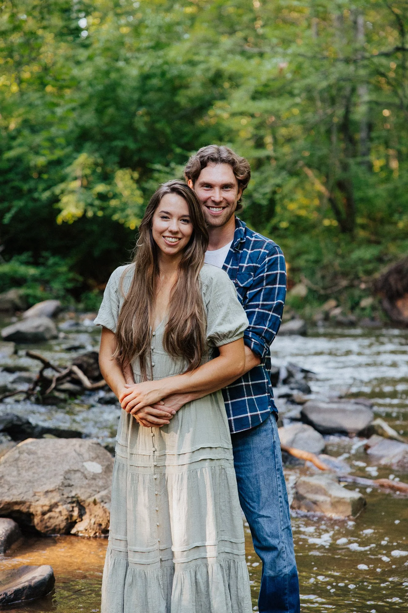 A smiling couple standing in a creek with rocks, surrounded by green trees in a forest.