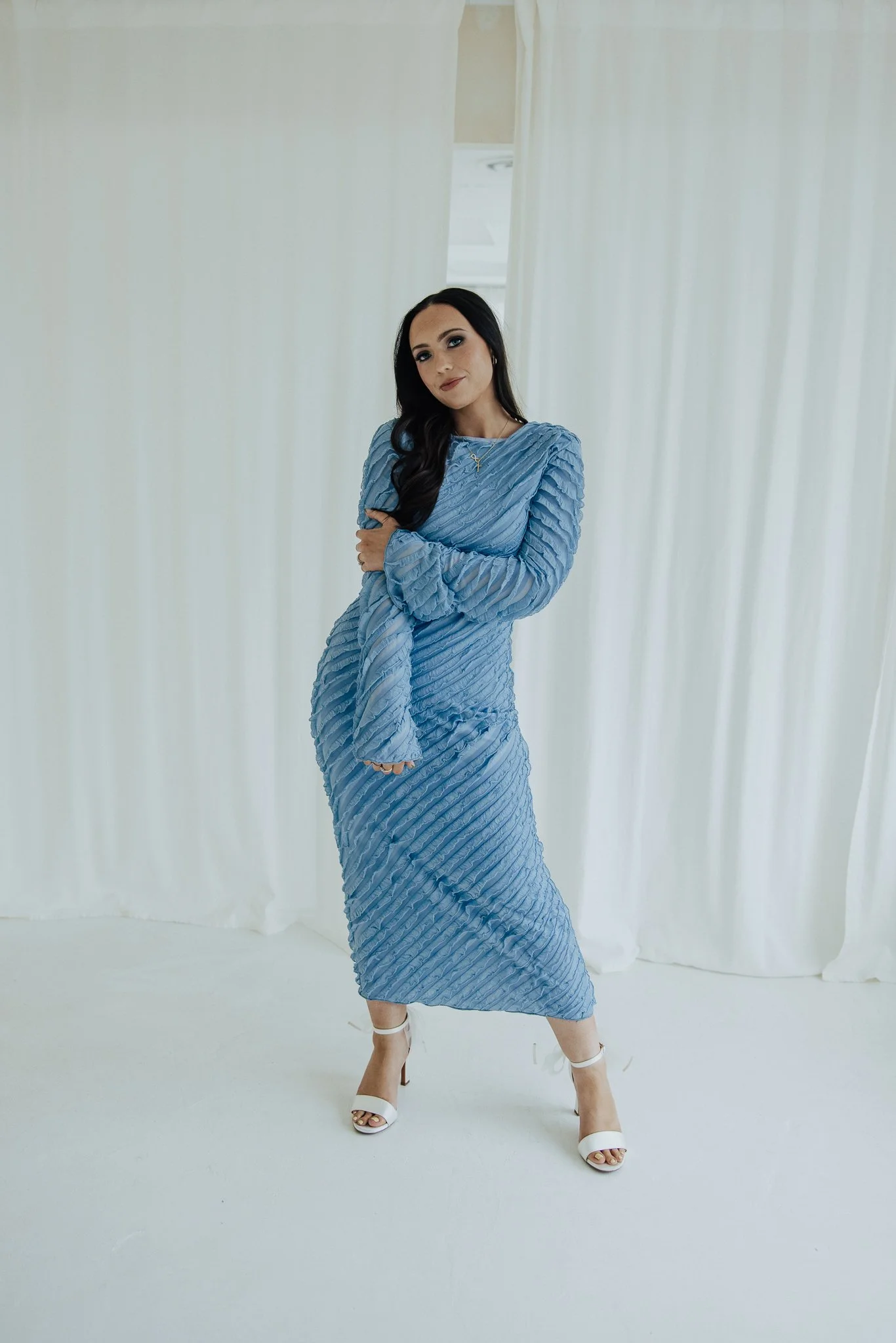 A woman in a blue textured dress and white high heels standing in front of white curtains, posing with her arms crossed and one leg slightly in front of the other.