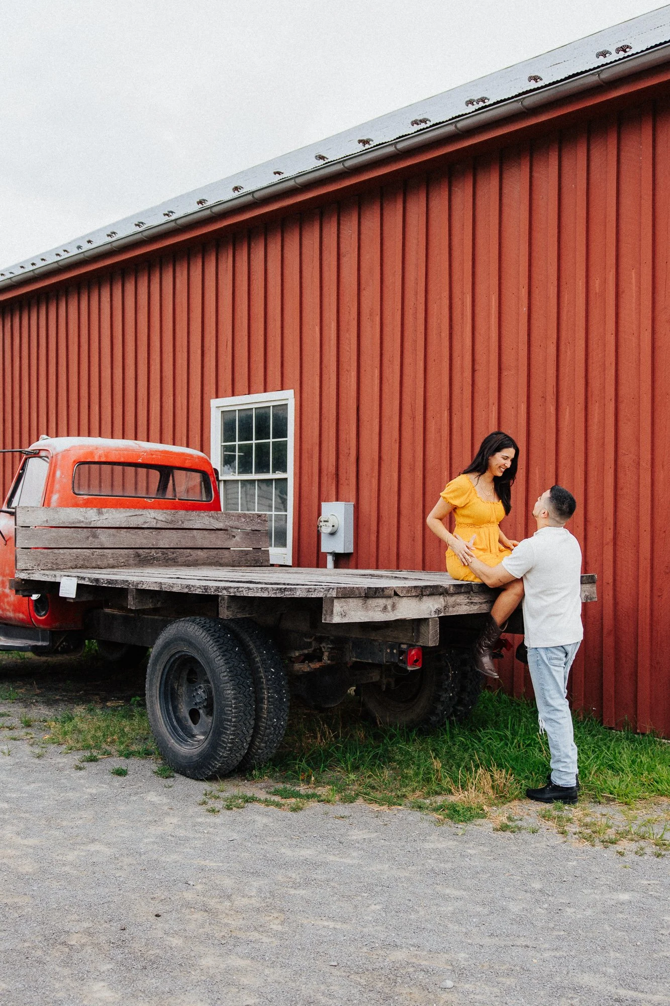 A woman in a yellow dress sitting on the back of a flatbed truck, holding hands with a man in a white shirt who is standing on the ground, in front of a red barn with a window, during daytime.