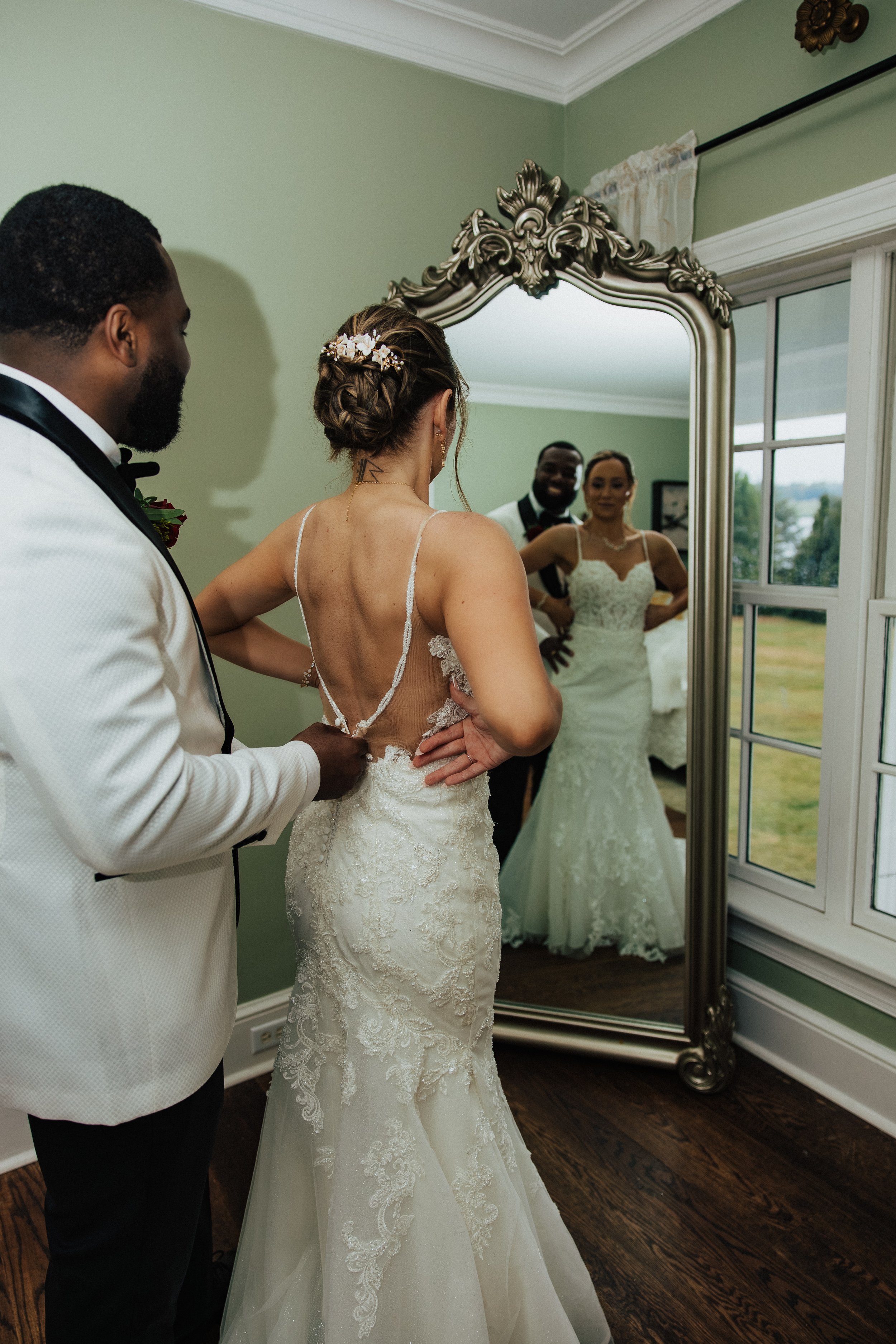 A bride and groom are looking at their reflection in a large decorative mirror inside a room with green walls and hardwood floors. The bride is adjusting her wedding dress, which has lace details, while the groom, dressed in a white tuxedo with black
