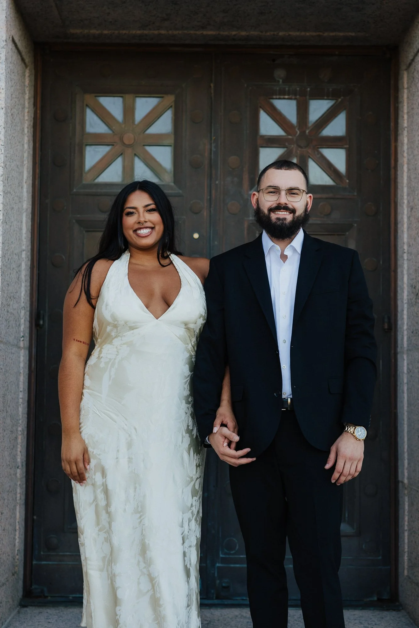 A smiling couple holding hands, standing in front of a large, ornate metal door. The woman is wearing a white dress, and the man is wearing a black suit and glasses.