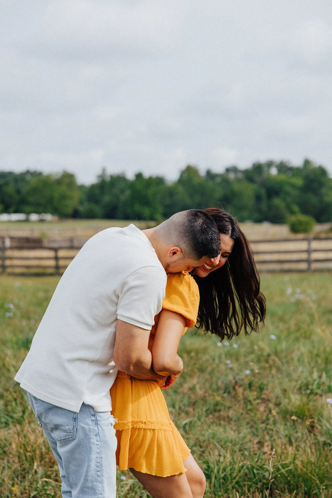 A young man and woman are laughing together outdoors in a grassy field, with a wooden fence and trees in the background. The man is wearing a white shirt and ripped jeans, and the woman is wearing a yellow dress.