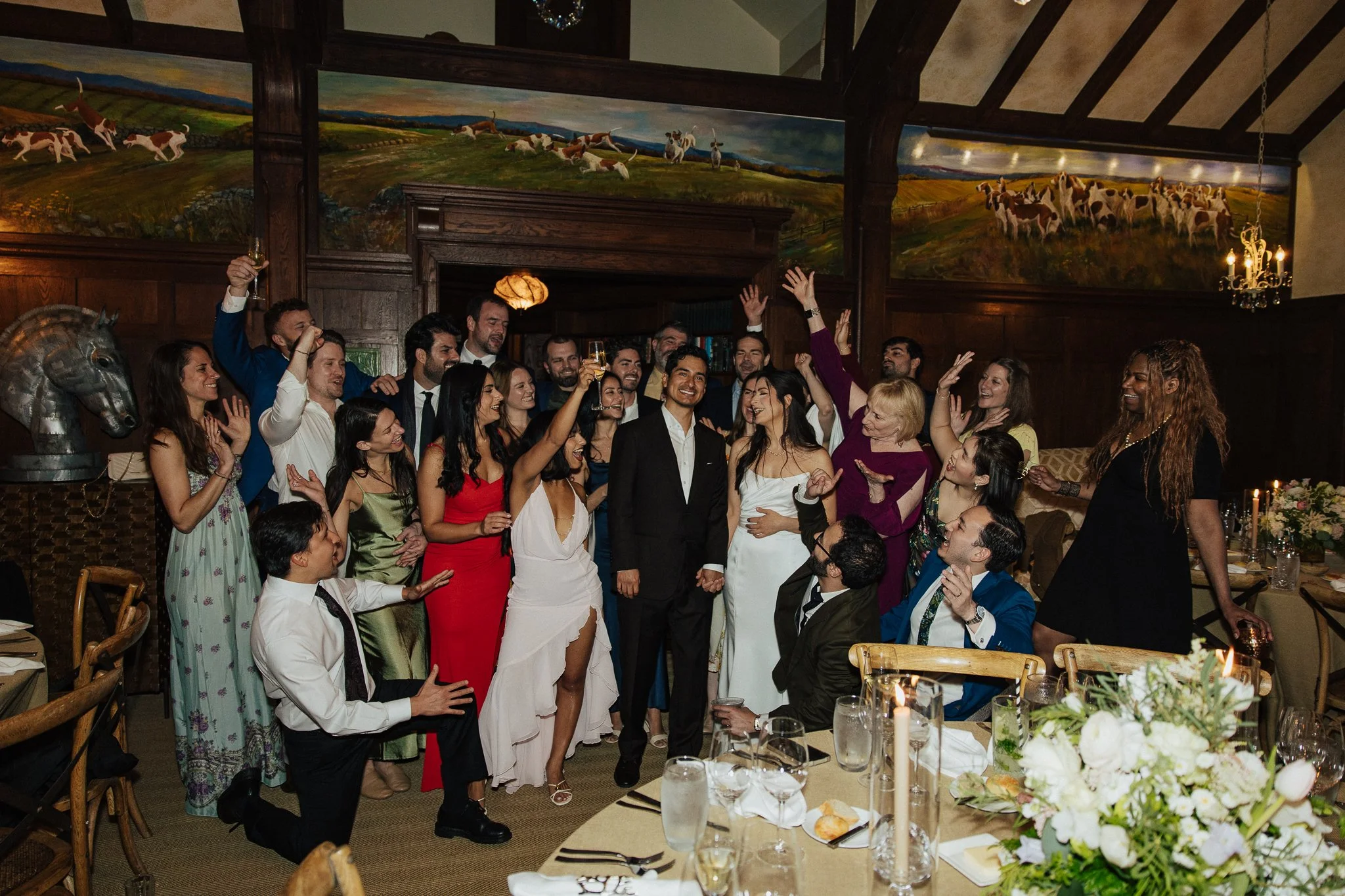 A large group of people celebrating at an indoor wedding reception, with a bride and groom in the center. The room is decorated with elegant wooden paneling, a colorful horse-themed mural on the wall, and floral centerpieces on tables.