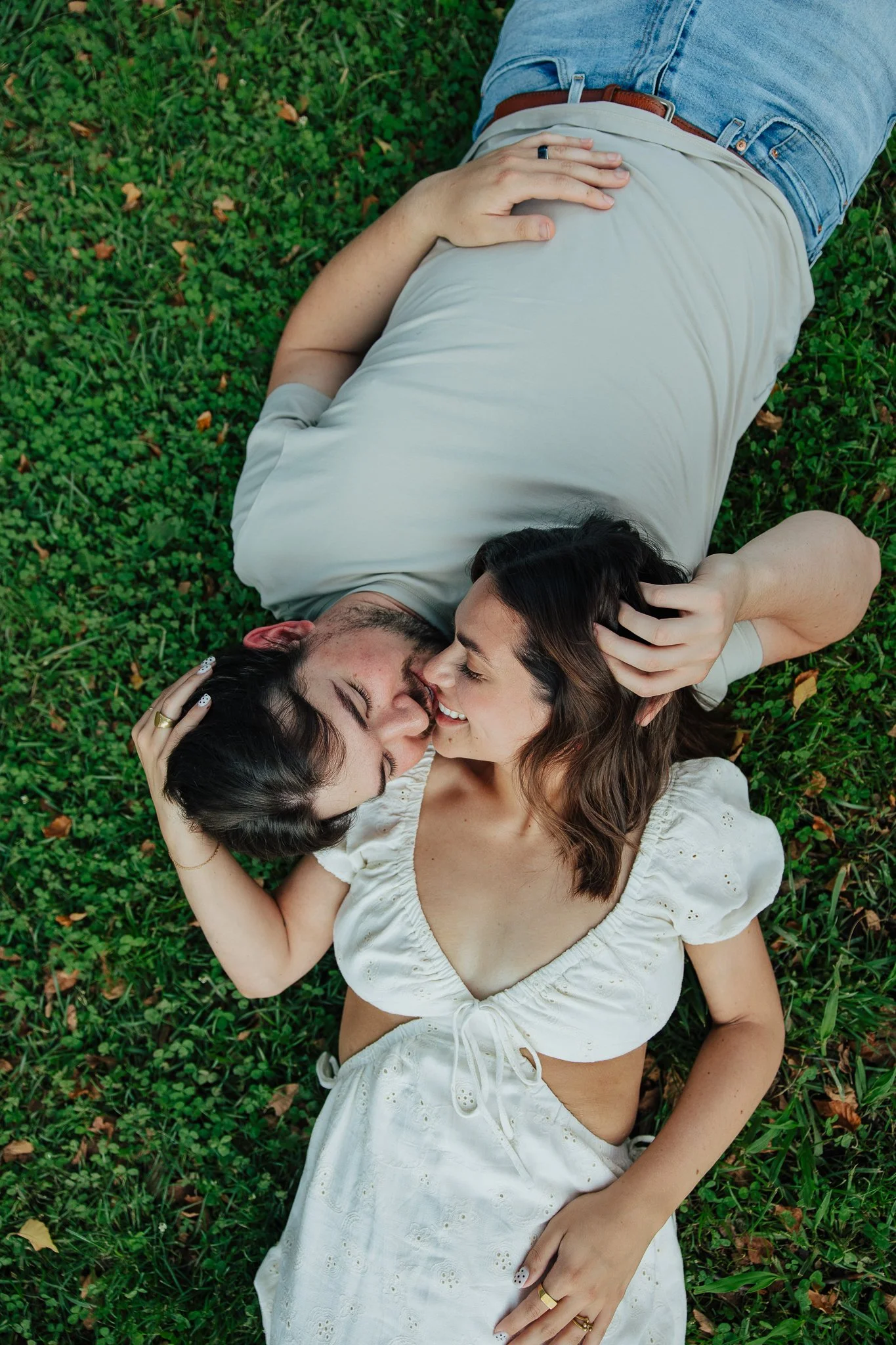 A man and woman lying on green grass, nose to nose, smiling and touching each other's faces.