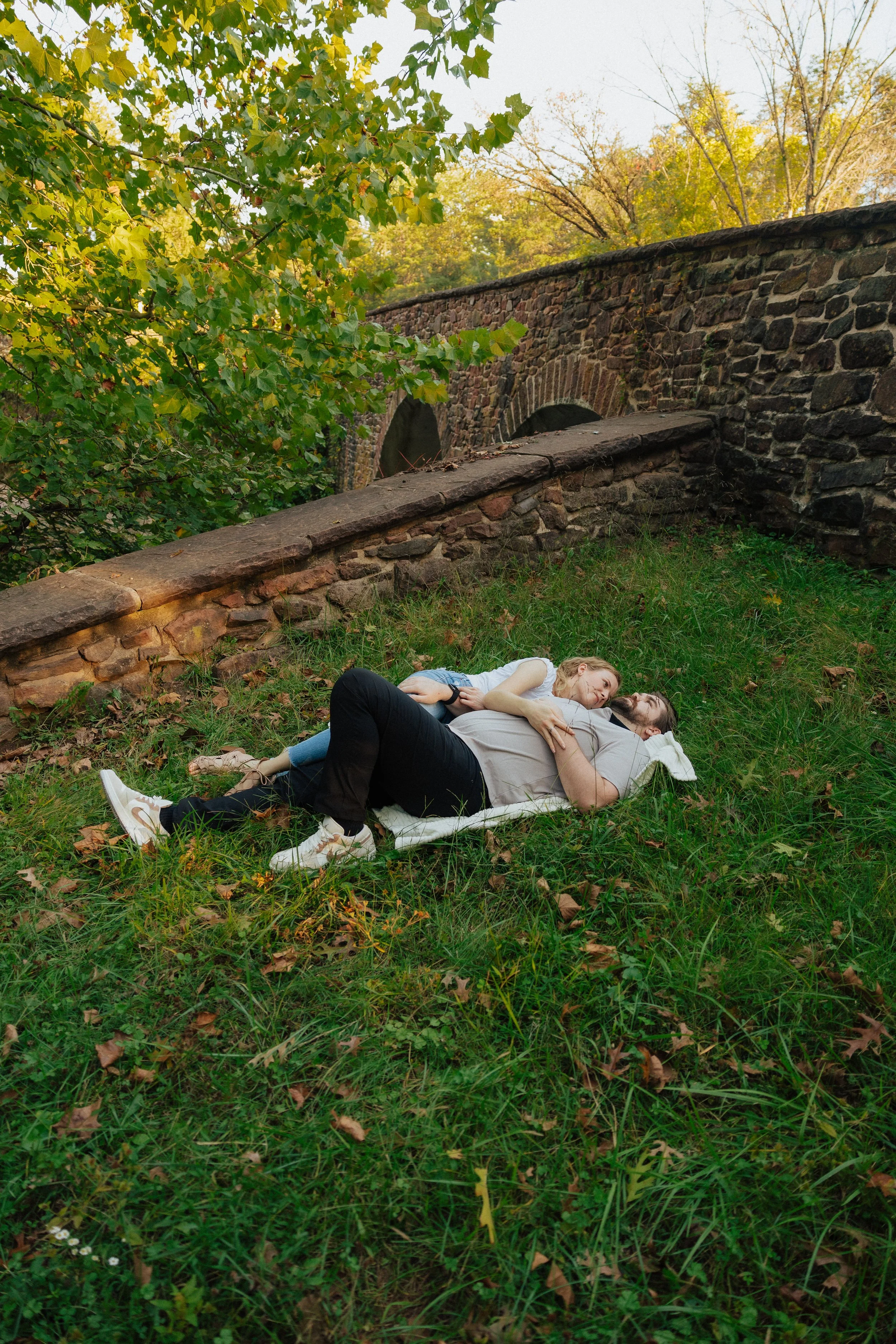 A couple lying on a blanket in a grassy area beneath trees, with a stone bridge in the background.