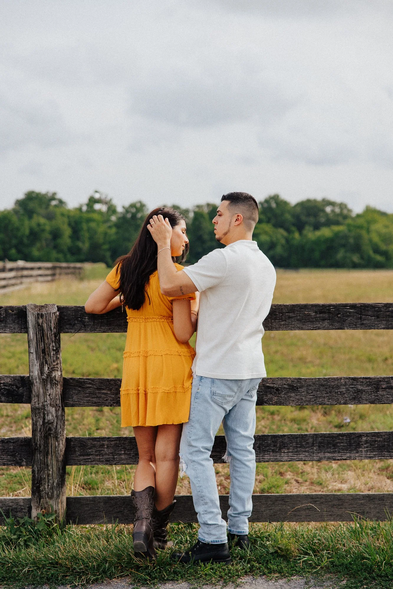 A young couple standing close together outdoors near a wooden fence, with trees and a cloudy sky in the background. The woman is wearing a yellow dress and boots, and the man is wearing a white shirt and light jeans.