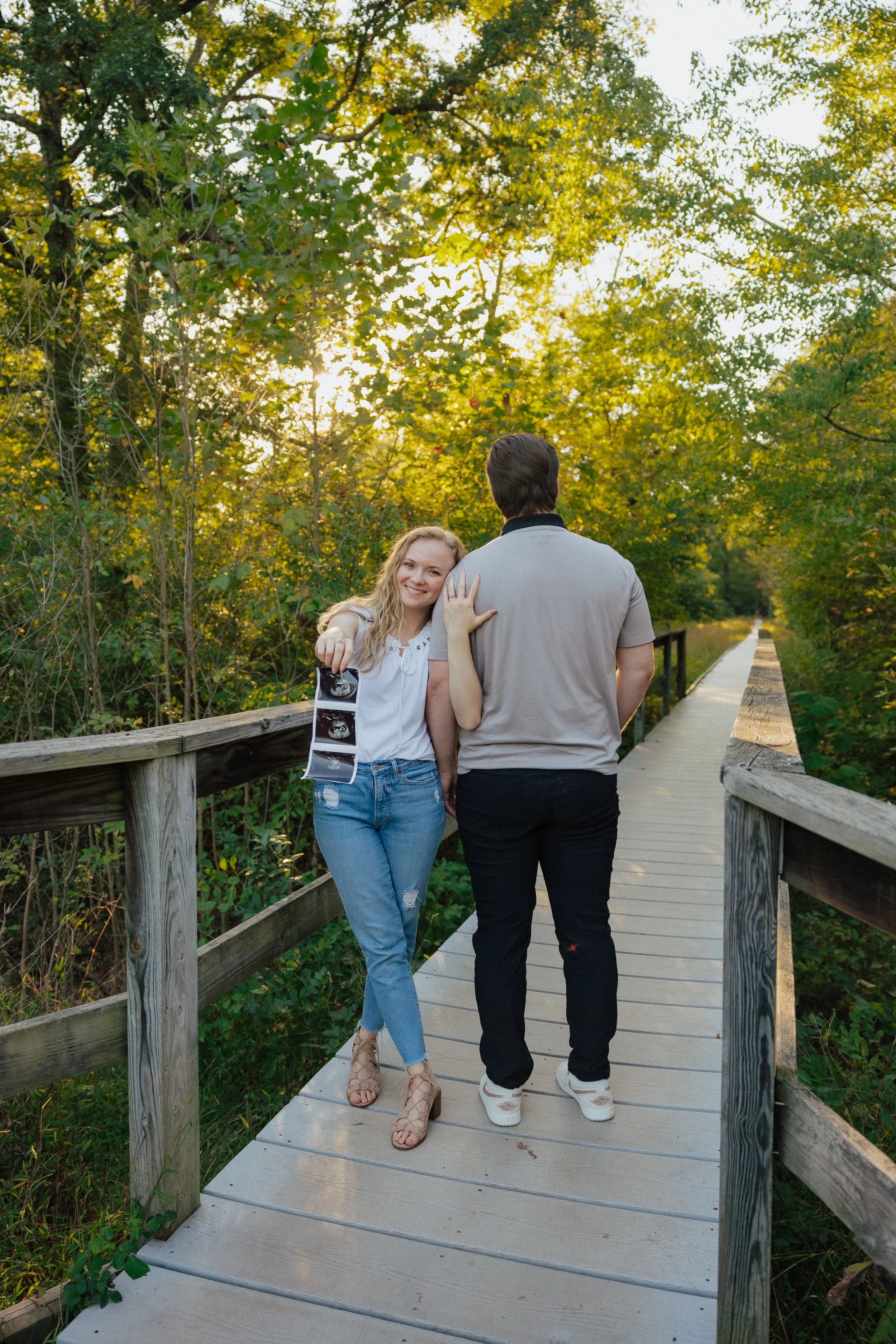 A woman and a man walking on a wooden bridge in a forest during the daytime. The woman is smiling and pointing towards the camera, holding a series of ultrasound images. The man is facing away from the camera, walking alongside her.