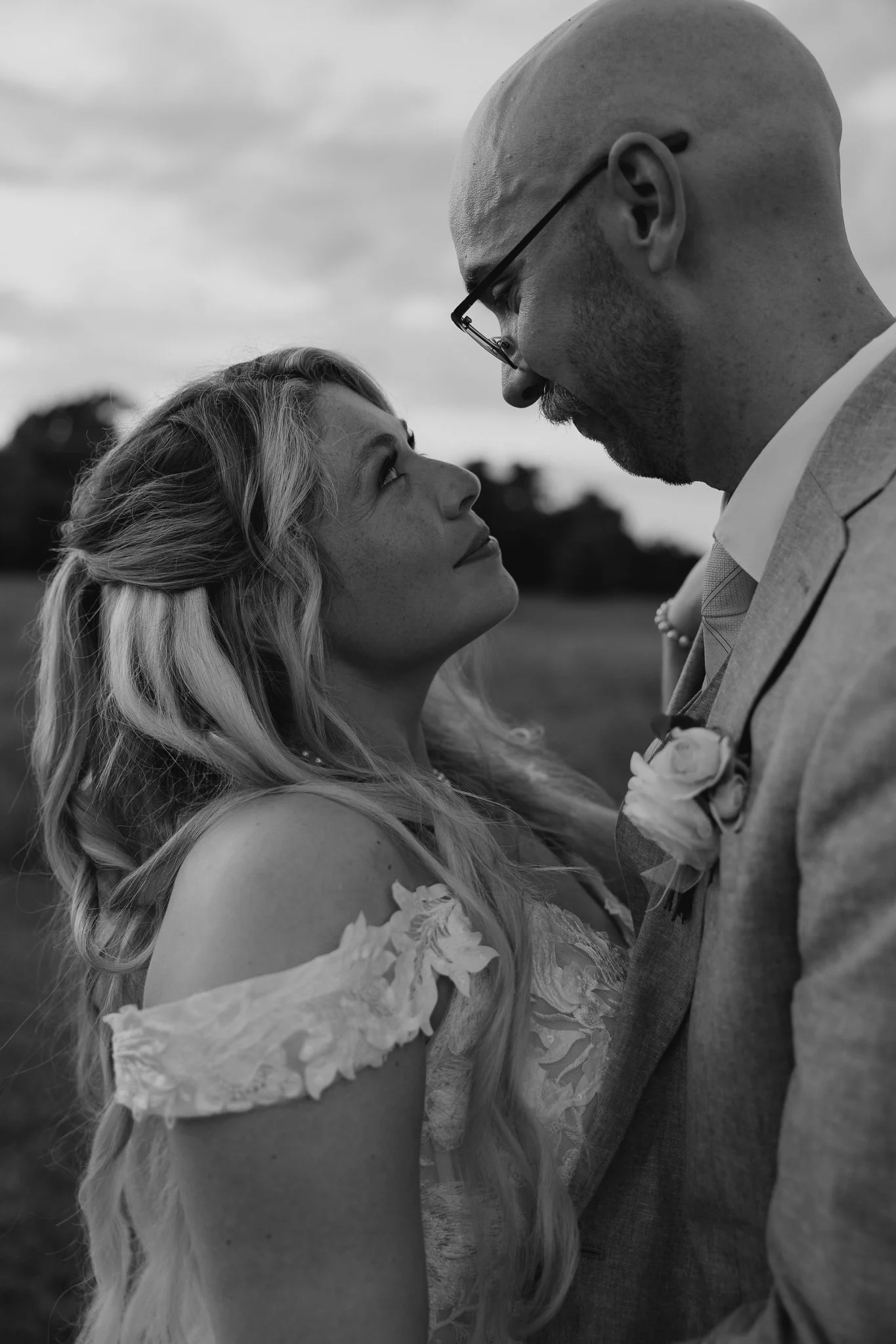 A black-and-white photograph of a couple facing each other closely outdoors, with the woman looking up at the man. The woman has long, wavy hair and is wearing a dress with lace details on the sleeves. The man is bald, wearing glasses, a suit, and a 
