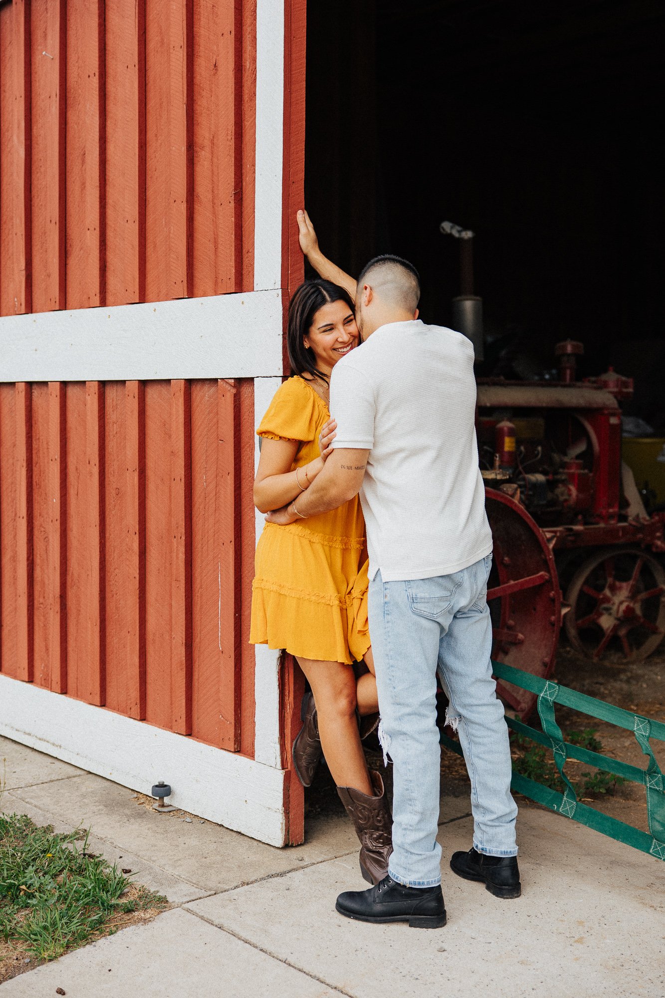 A man and woman embrace by a red barn, with a farm equipment in the background, smiling and looking at each other.