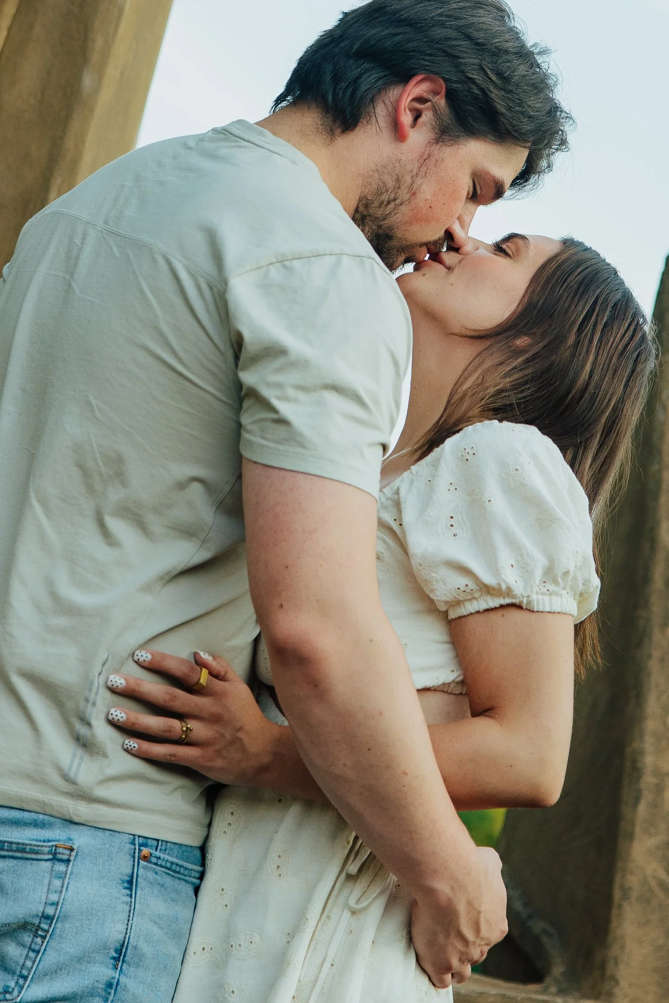 A man and a woman are kissing closely outdoors, with the man leaning down towards the woman, who has her eyes closed and head tilted back. The image captures an intimate moment between the two.