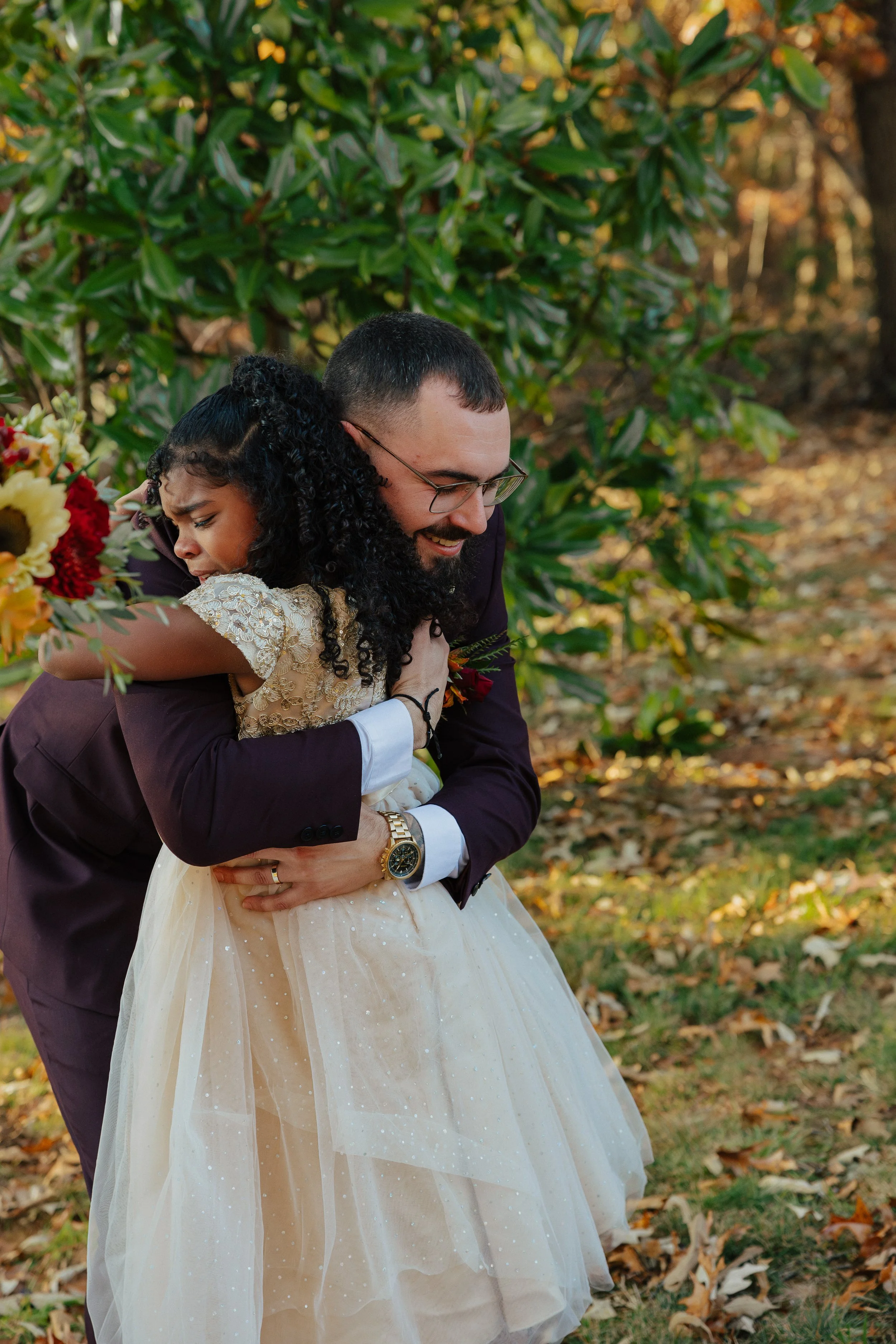A groom hugging a young girl in a forest with fallen leaves, during autumn, with trees and sunlight in the background.