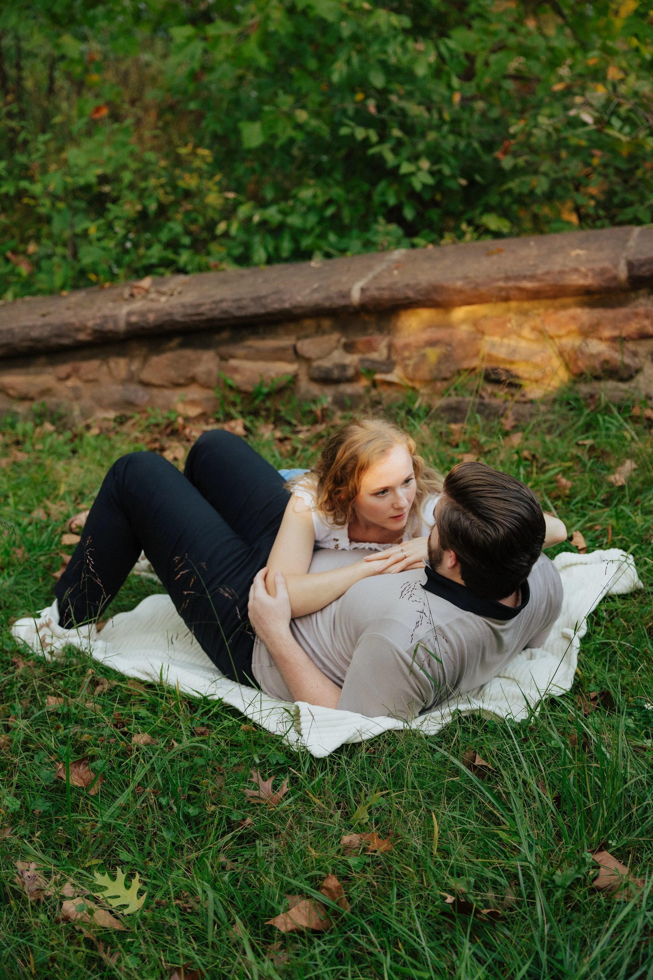 A young woman and man lie on a white blanket in a grassy outdoor area, facing each other with their heads close together. The woman has curly blonde hair and is looking at the man, who has dark hair and glasses. There is a stone wall and greenery in 