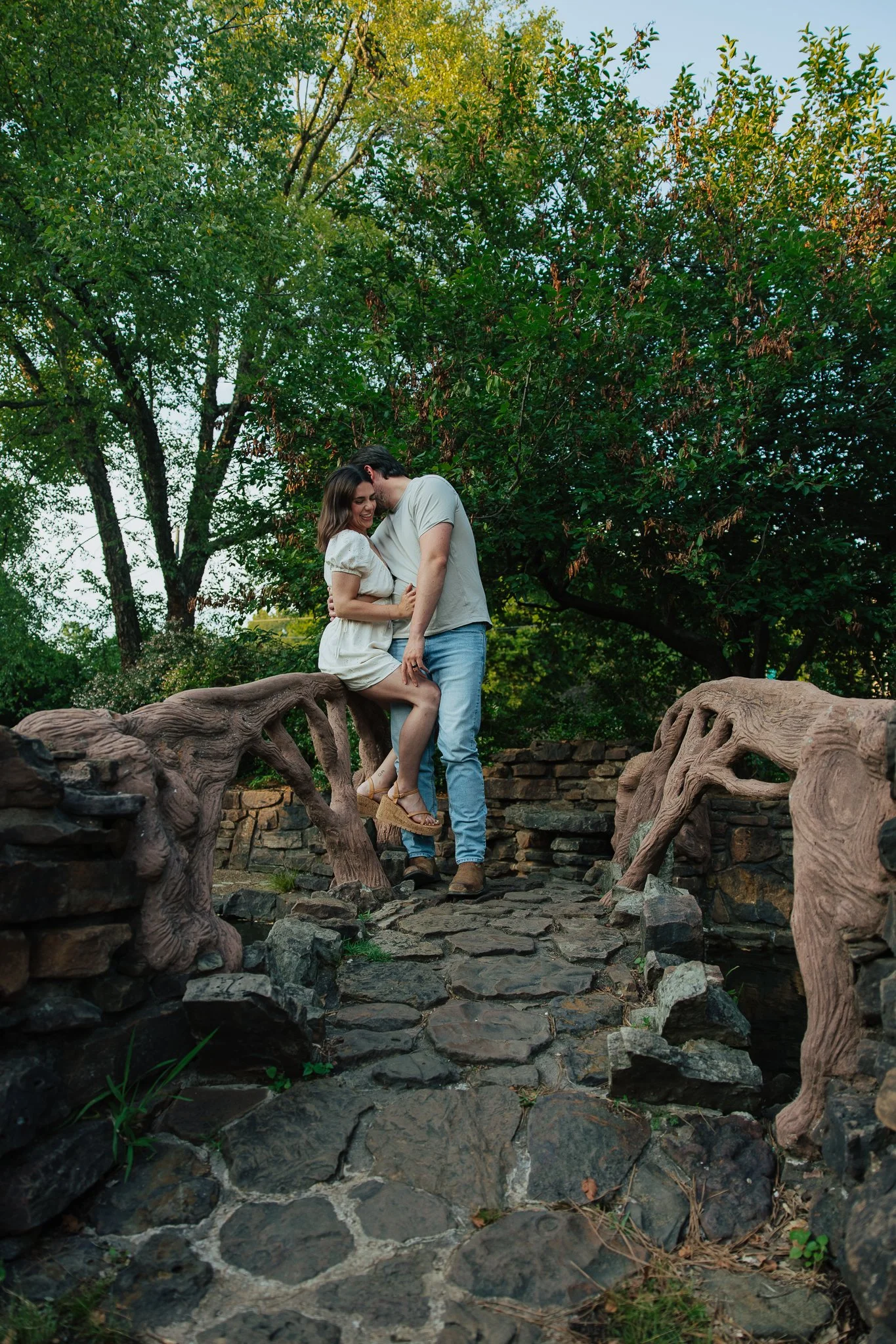 A couple stands close together on a decorative small bridge with carved wooden railings over a rocky stream. They are smiling, facing each other, with the woman's hands on his chest. Behind them is lush green foliage with tall trees, creating a natur