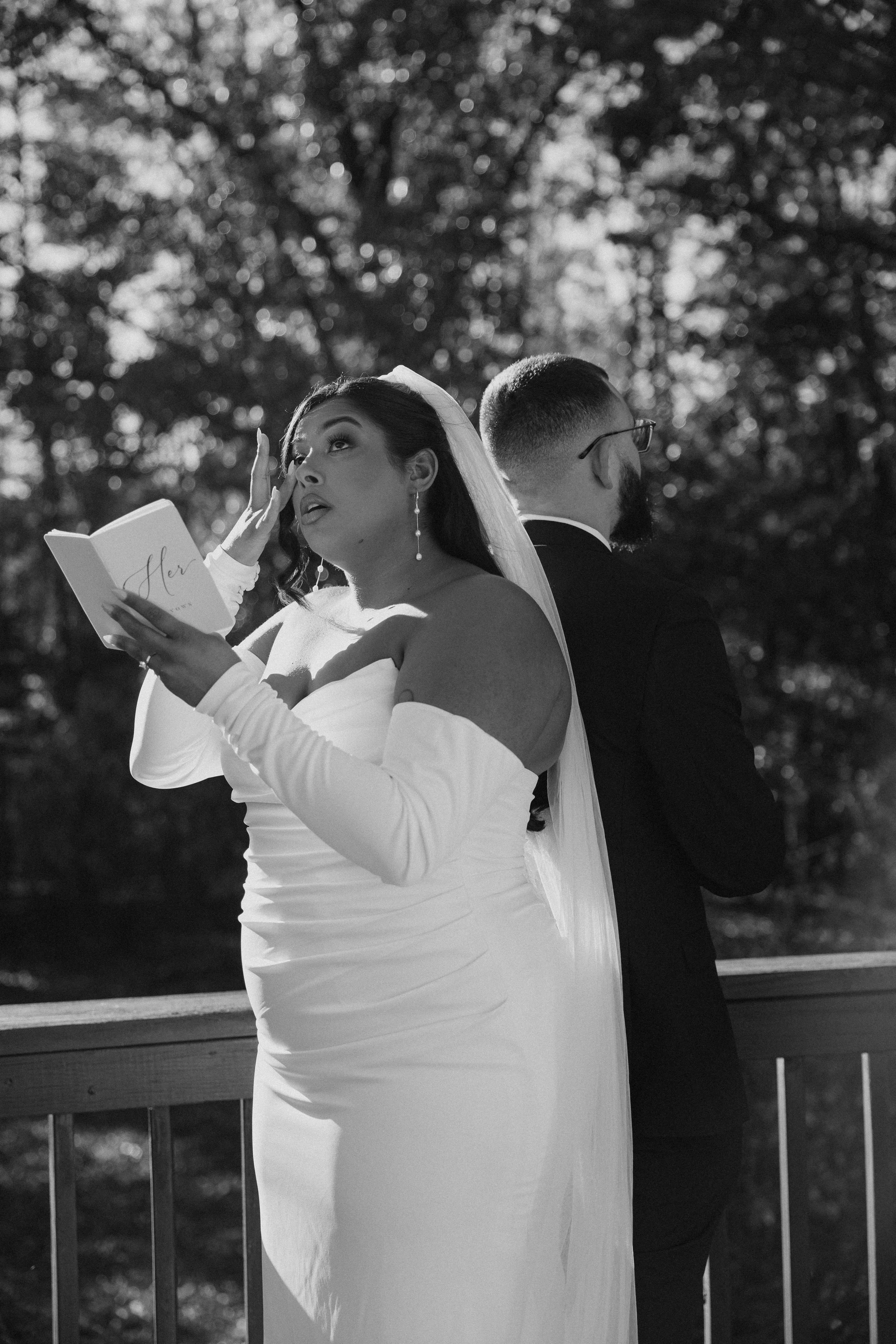 A woman in a wedding dress and a man in a suit stand back to back outdoors, with trees in the background. The woman appears emotional, holding a small book or card, while the man has his back turned, wearing glasses.