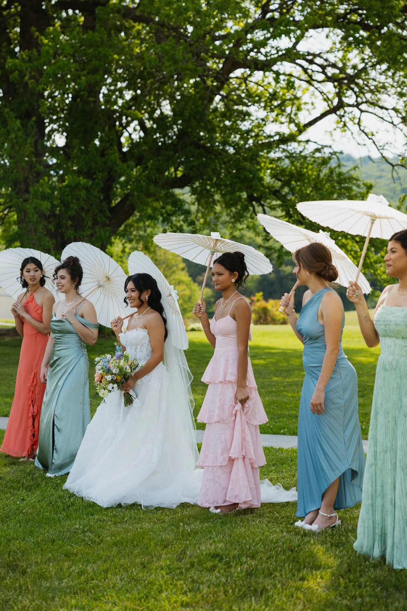 A group of women in formal dresses holding white parasols, standing outdoors on green grass with large trees in the background, possibly at a wedding or special event.