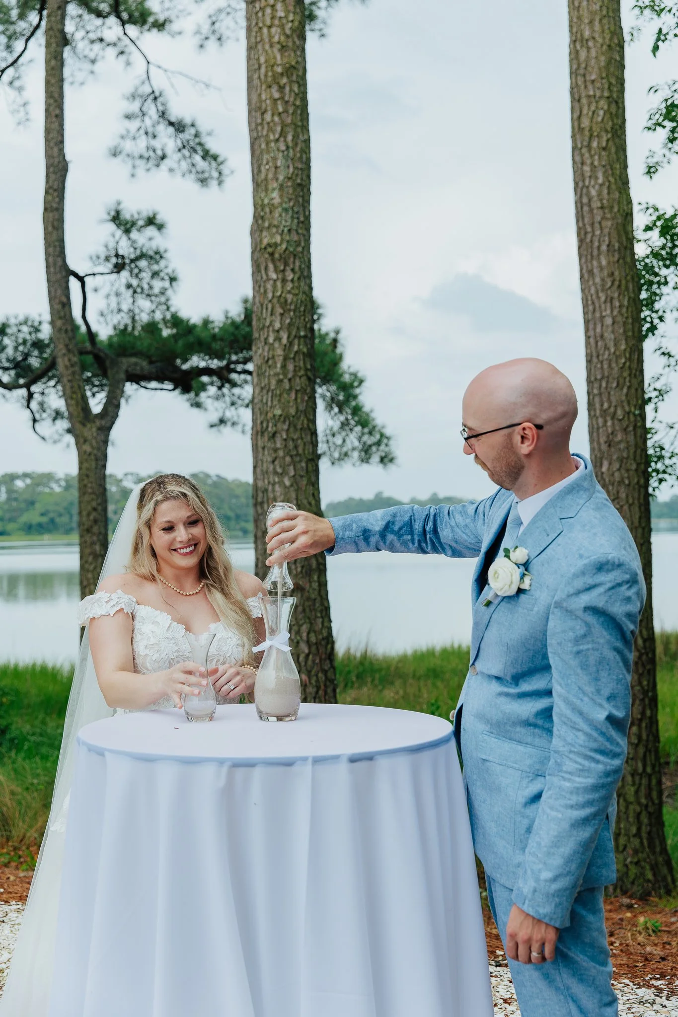A wedding ceremony outdoors with a bride and groom. The groom, wearing a light blue suit, is pouring wine into a glass for the bride, who is smiling and holding a glass. They stand near a round table with a white tablecloth, in front of a lake and ta