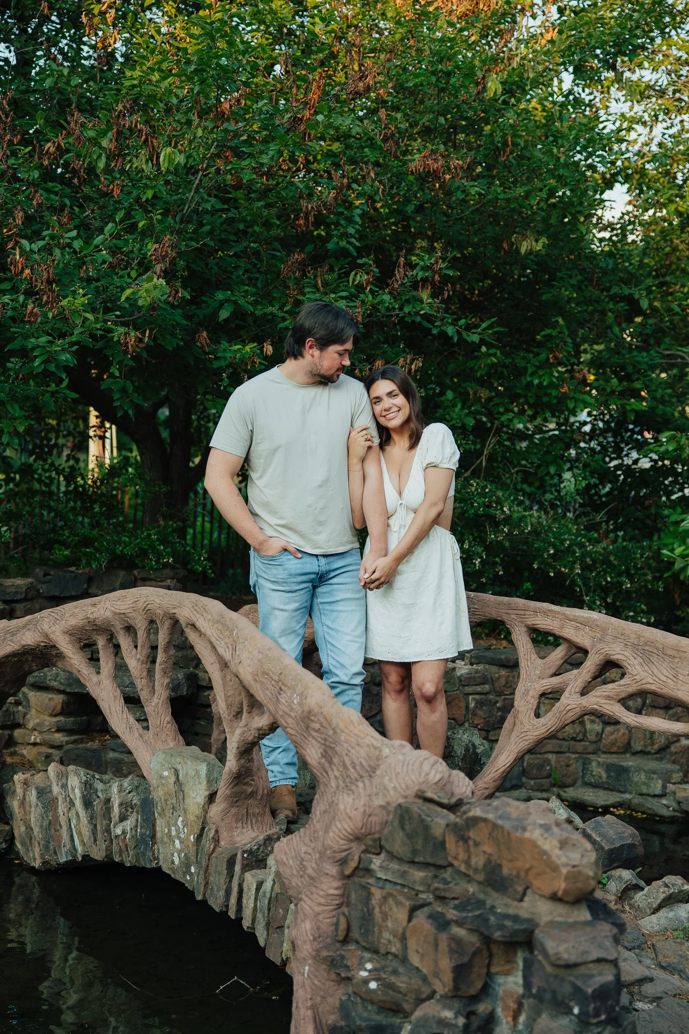 A man and woman standing on a stone and wood bridge above water, holding hands, with green trees in the background.