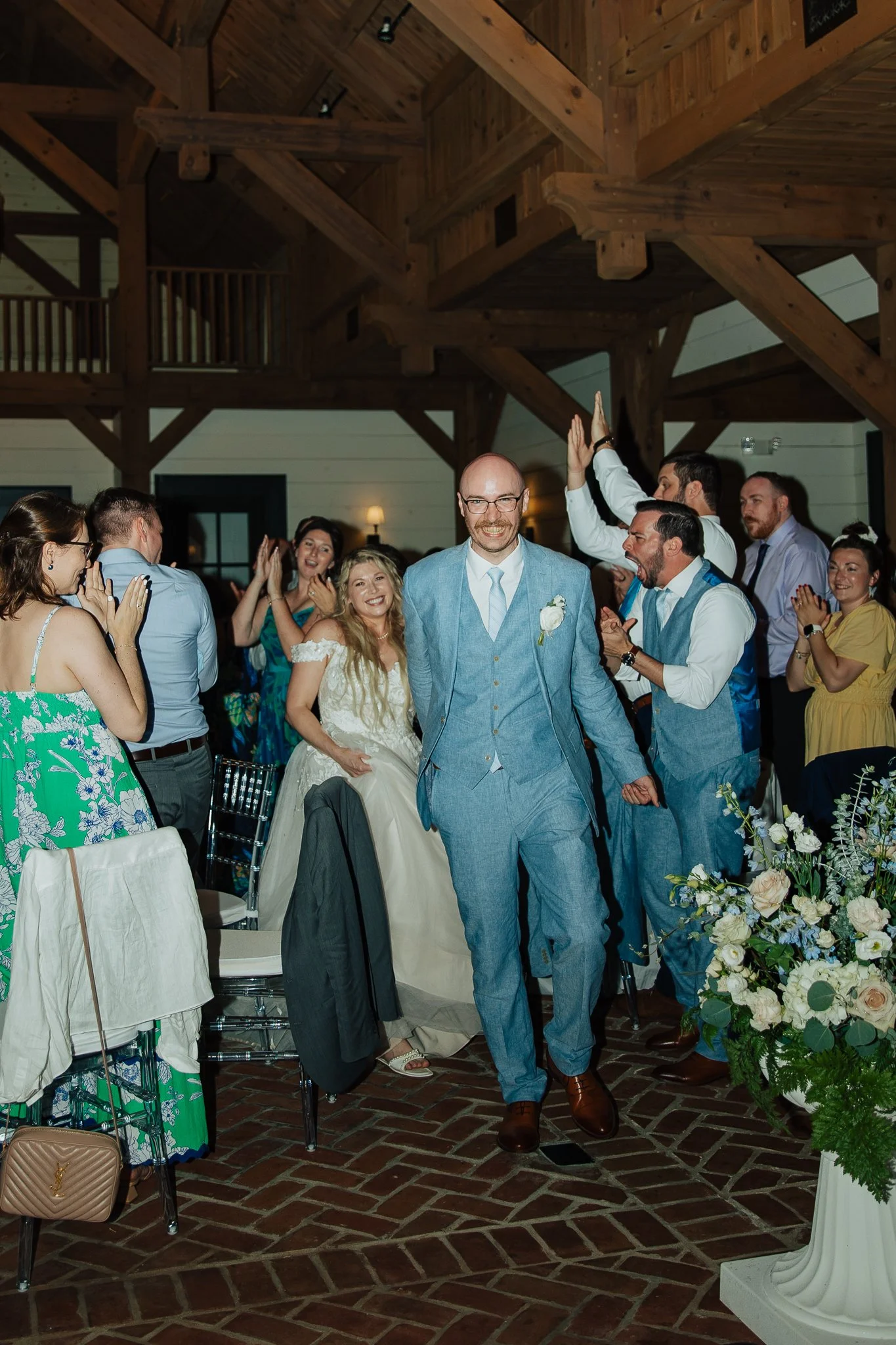 People celebrating at a wedding reception with a groom in a blue suit and a bride in a white gown surrounded by guests in an indoor rustic venue with wooden beams and floral arrangements.