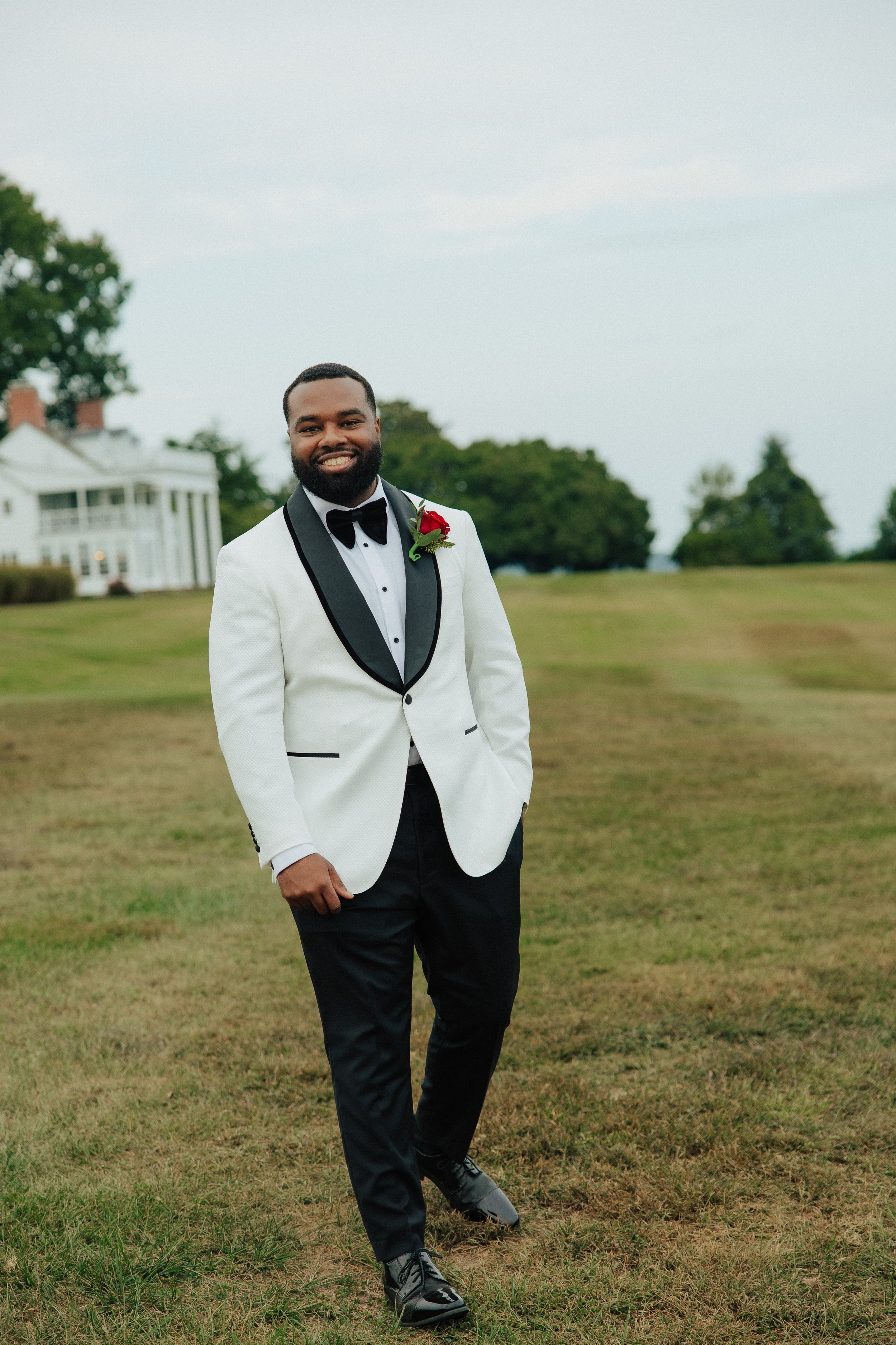 A man in a tuxedo with a red boutonniere stands outdoors on grass, smiling at the camera with a large house and trees in the background.