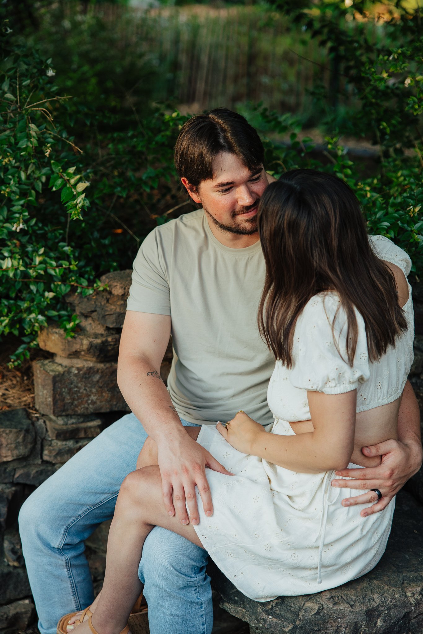 A couple sitting outdoors on a rock surrounded by greenery, sharing a tender moment. The man is wearing a light-colored T-shirt and jeans, and the woman is in a white dress with puffed sleeves.