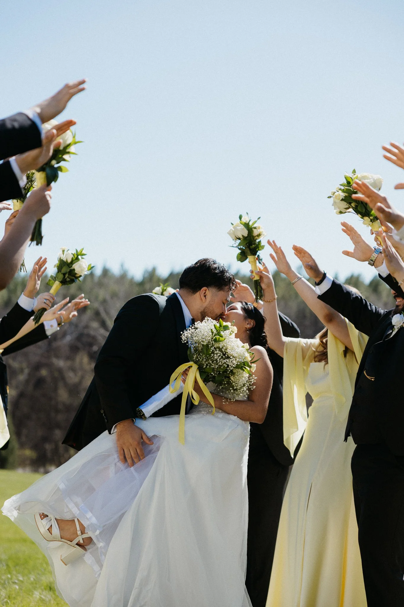 A bride and groom kissing outdoors during their wedding, surrounded by guests holding bouquets and raising their hands in celebration.