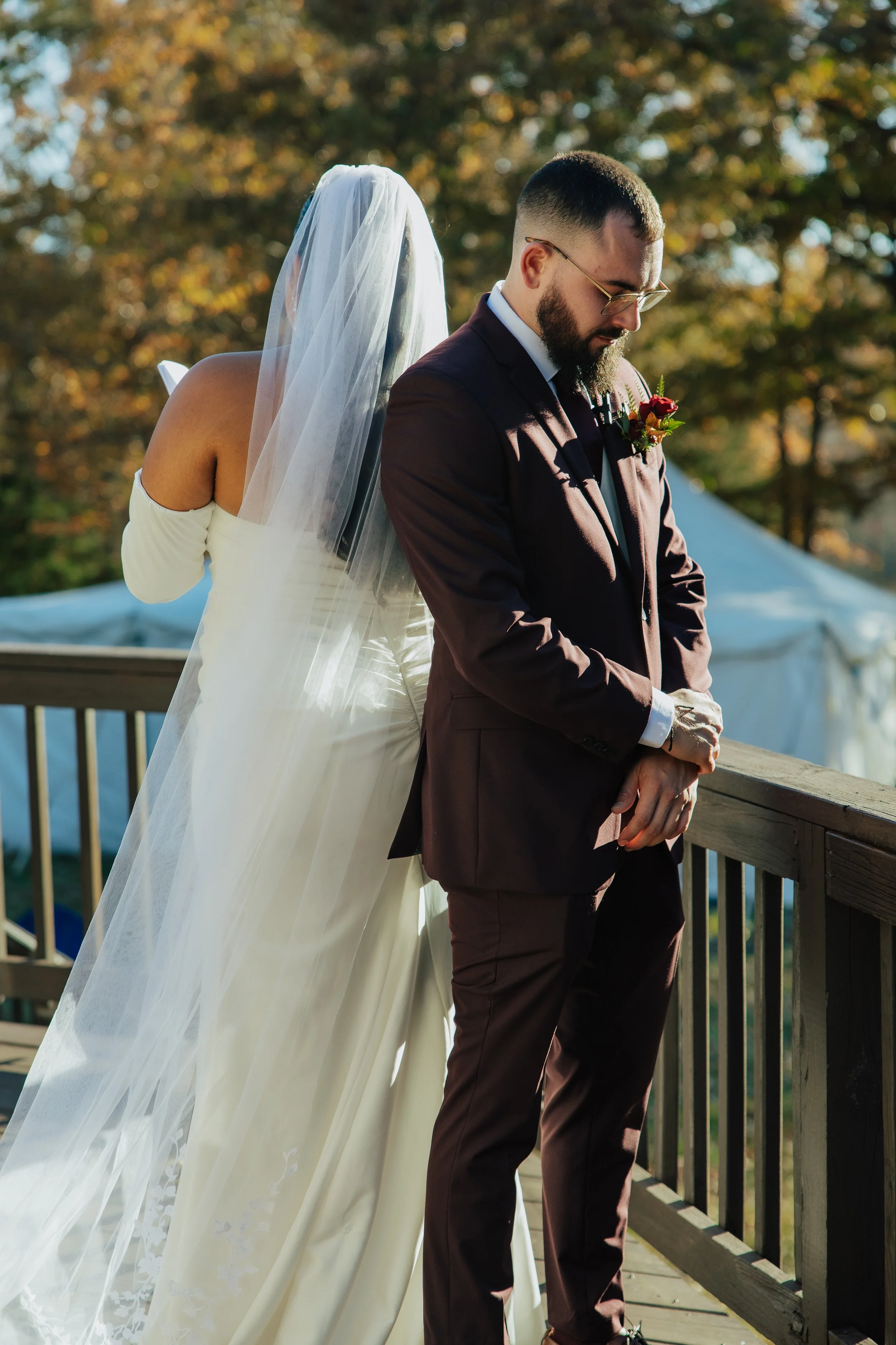 A bride and groom standing on a wooden deck during a wedding ceremony. The bride is wearing a white off-the-shoulder wedding dress with a long veil, and the groom is dressed in a dark suit with a boutonniere. They appear to be praying or in a moment 