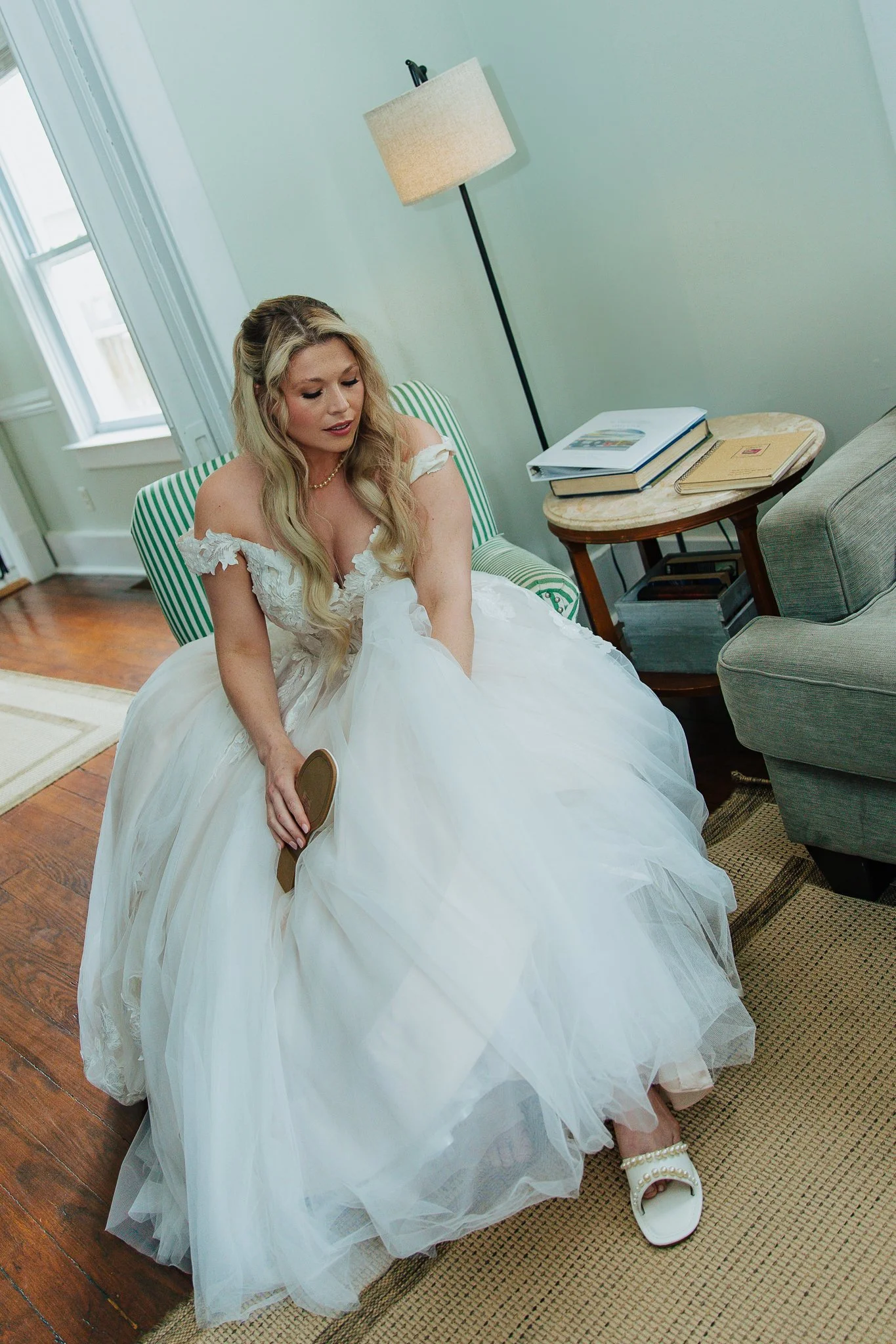 A woman in a white wedding dress sitting on a striped green and white chair, holding a shoe, with her left foot partially visible wearing a white sandal with pearls, in a room with a window and a side table with books and a magazine.