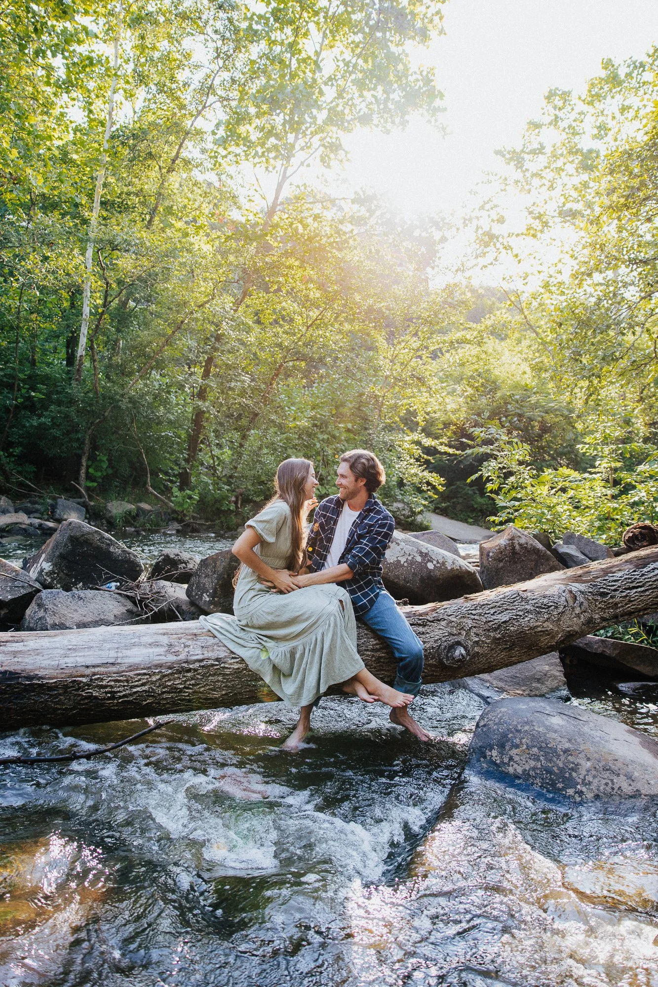 A couple sitting on a fallen tree trunk in a stream, surrounded by green trees, with sunlight shining through the leaves.