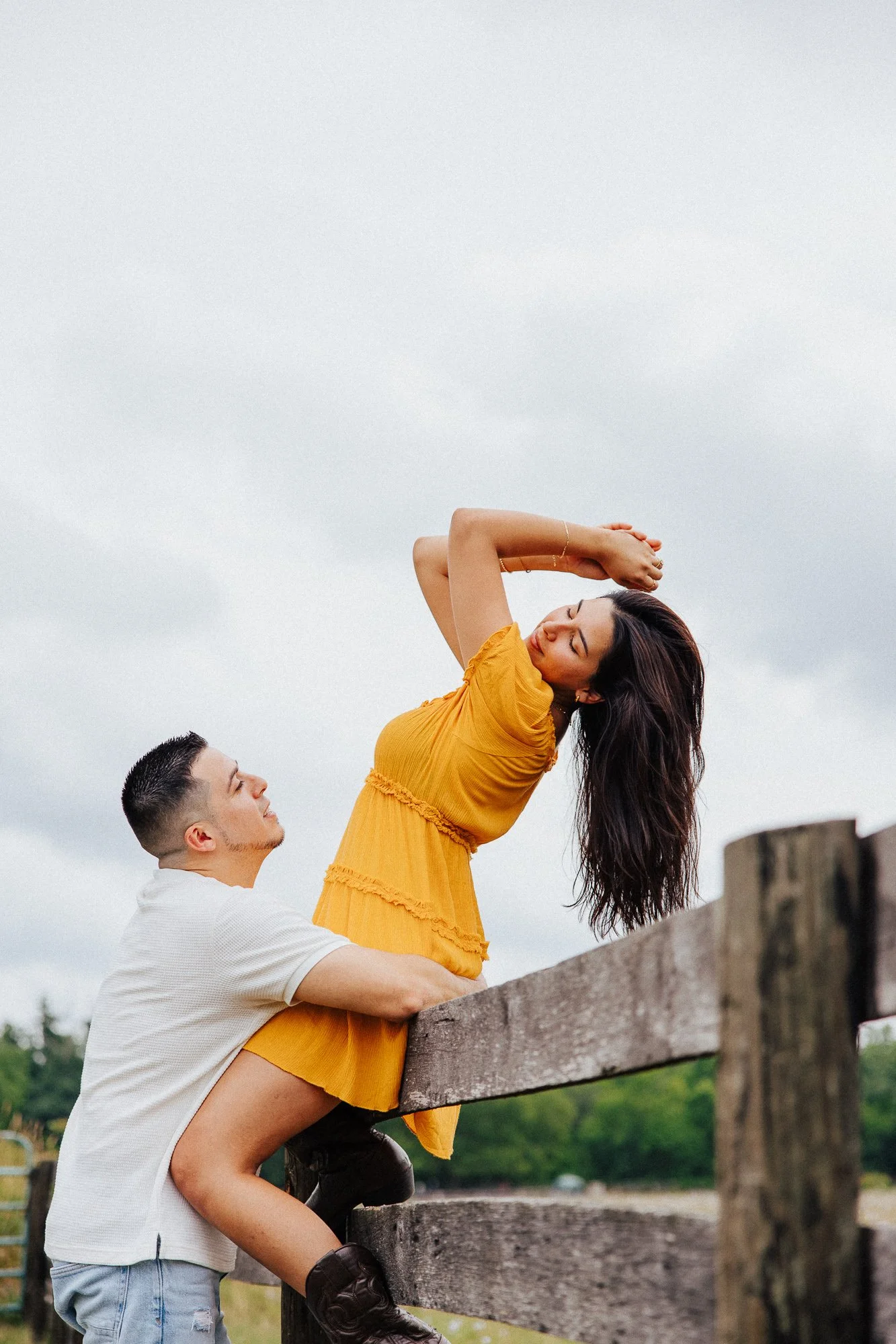 A man lifts a woman wearing a yellow dress onto a wooden fence outdoors. The woman has long dark hair and is holding her arms above her head while the man, dressed in a white shirt, supports her from below. The sky is cloudy and there are trees in th