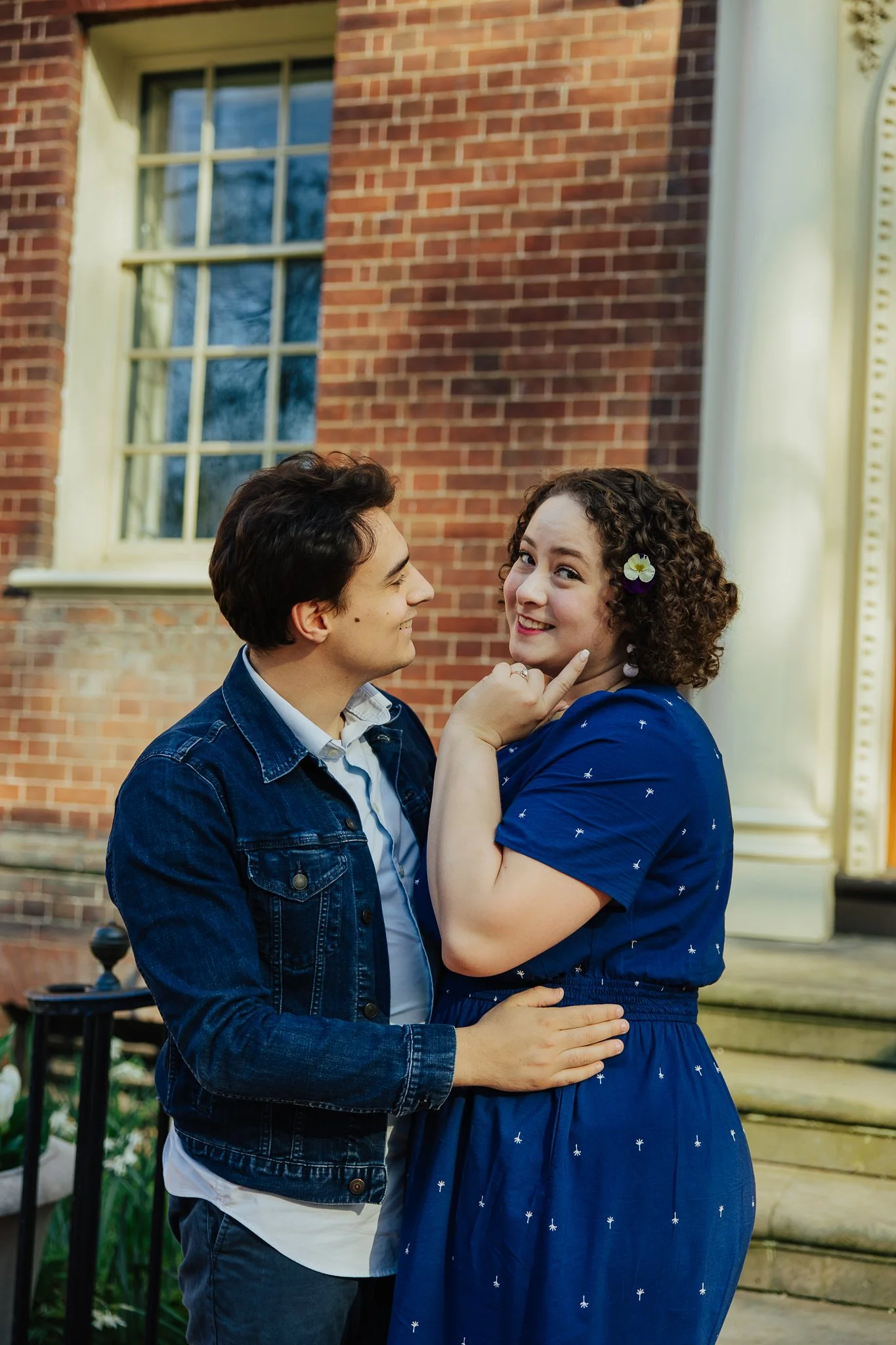 A young man and woman are in a close, playful pose outside a brick building. The woman has curly hair with a small flower hair accessory and is smiling while touching the man's face. The man is looking at her with a smile, wearing a denim jacket.