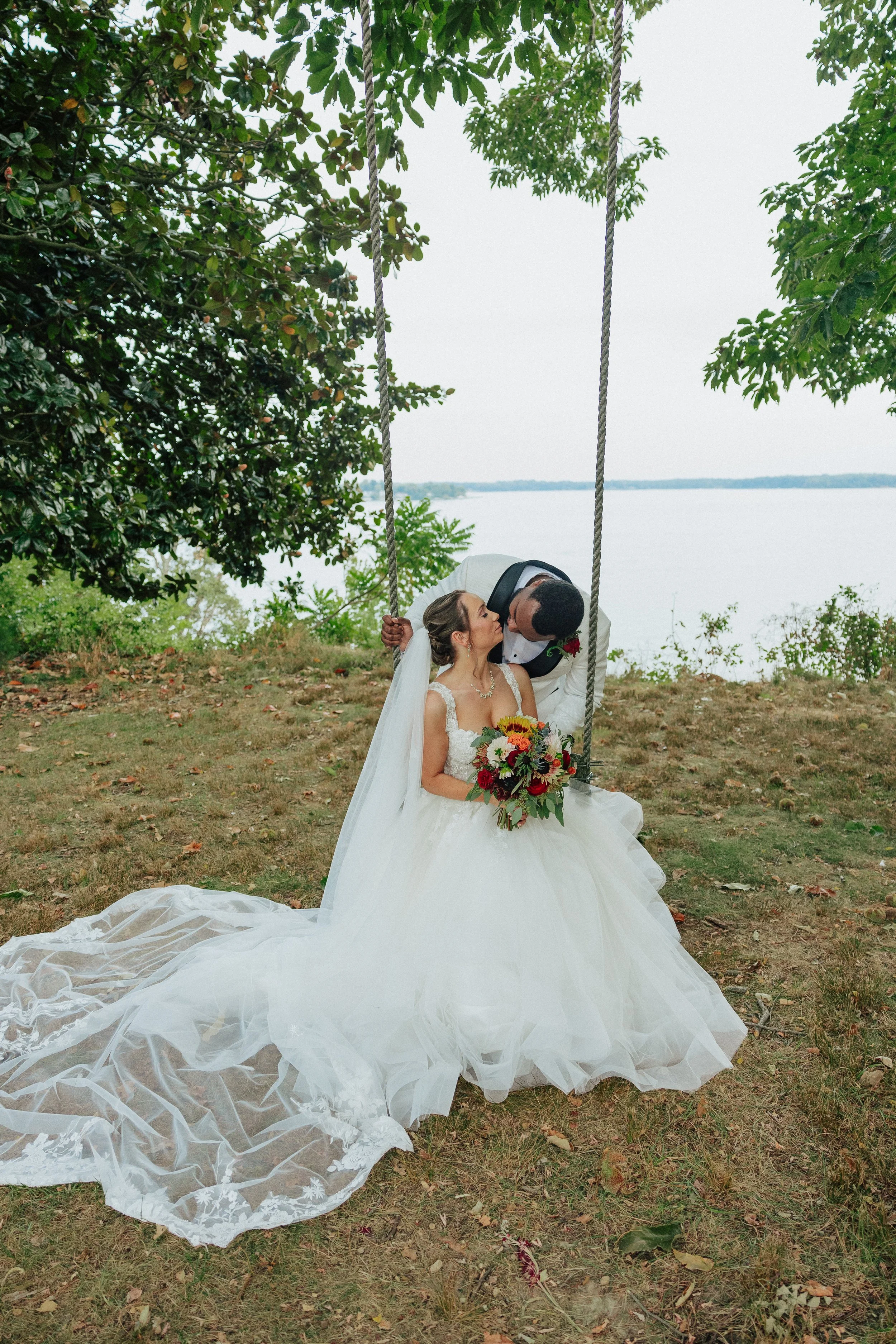 A bride and groom sharing a kiss outdoors near a lake, with the bride sitting on a swing, holding a bouquet of flowers, and the bride in a white wedding dress with a long train.