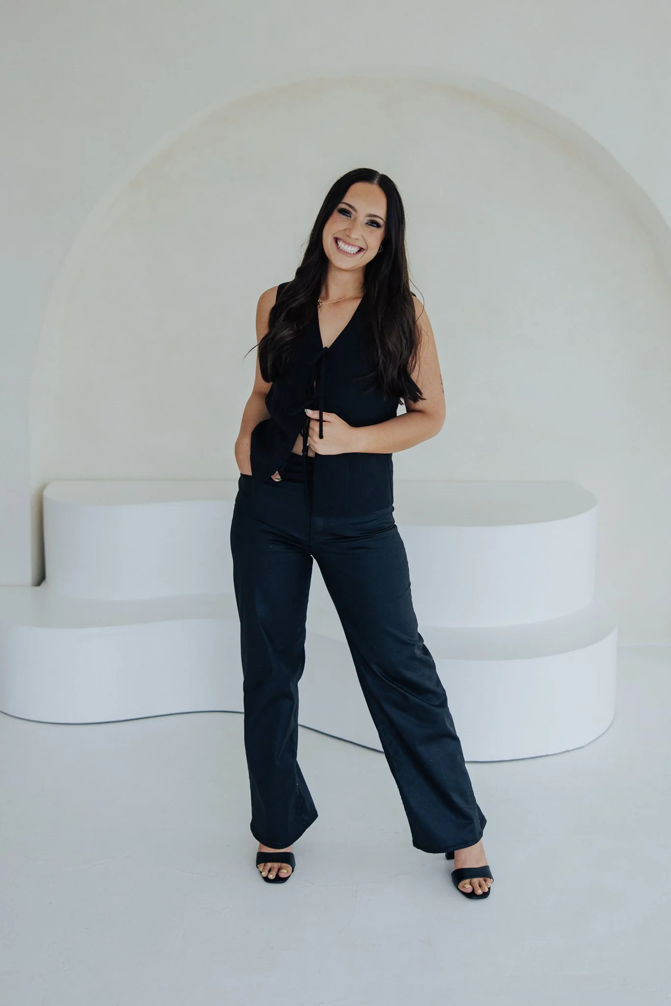 Young woman with long dark hair smiling and posing in a black sleeveless top and black pants, standing in front of a white abstract wall with a circular design