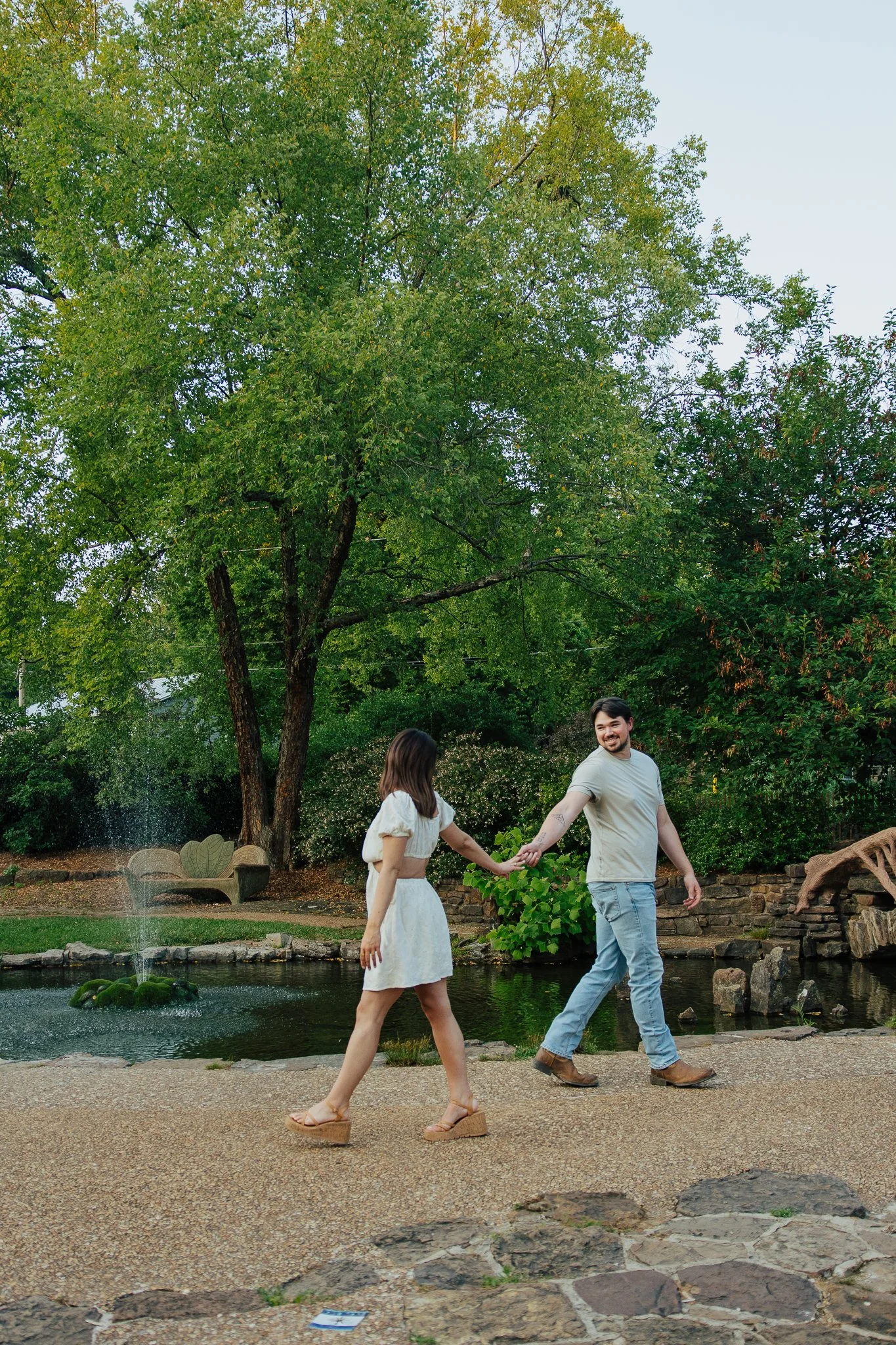 A man and a woman holding hands and walking near a pond in a park-like setting with large trees, a decorative bench, rocks, and a small fountain in the background.