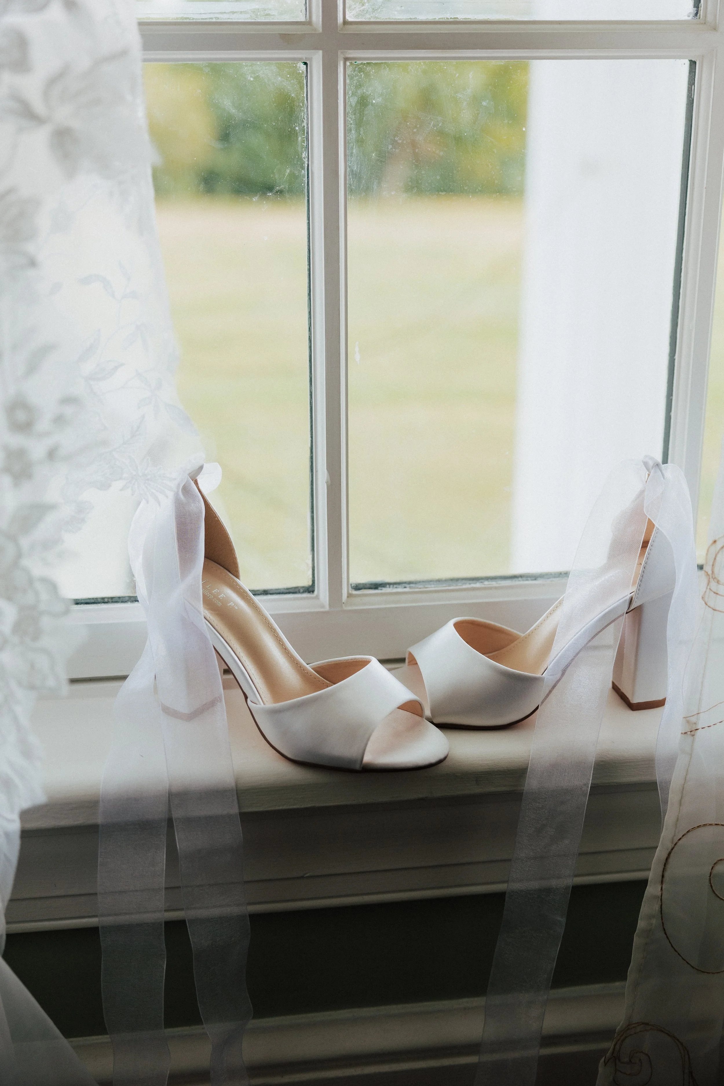 A pair of white high-heeled shoes with ankle ribbons and a pair of matching white shoes placed on a windowsill. The window has a white frame and sheer curtains with a floral pattern, letting in natural light.