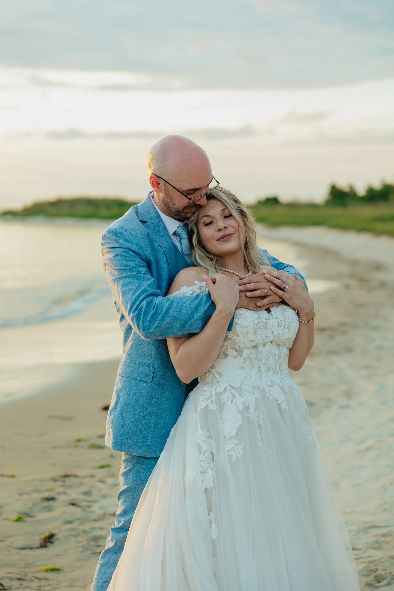 A newlywed couple embraces on a beach with the ocean and cloudy sky in the background. The groom is in a blue suit and the bride is in a white wedding gown with lace details, wearing a pearl necklace.