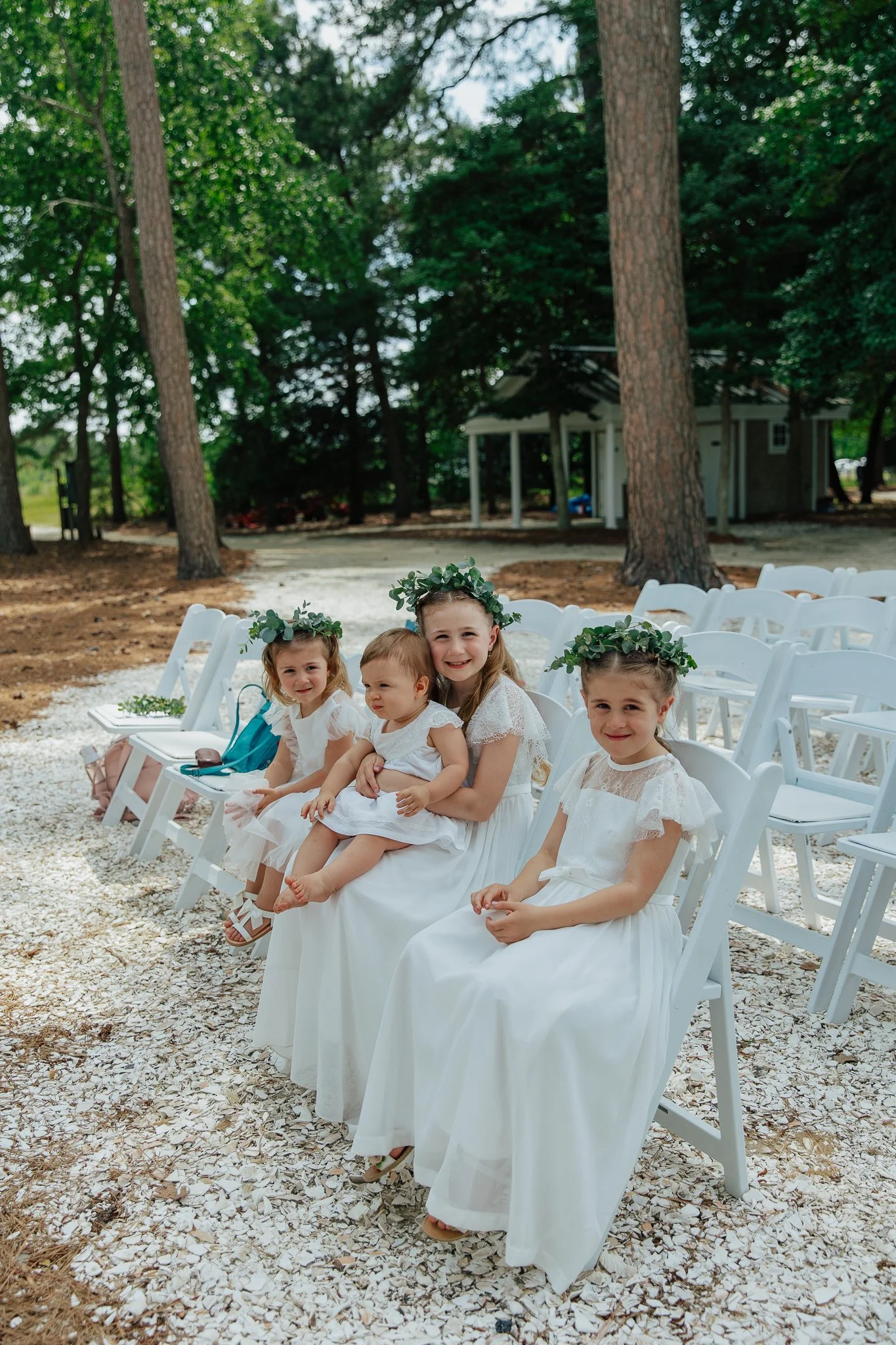 A group of young girls dressed in white dresses with flower crowns, seated outdoors on white chairs at a gathering, surrounded by trees and a small building in the background.