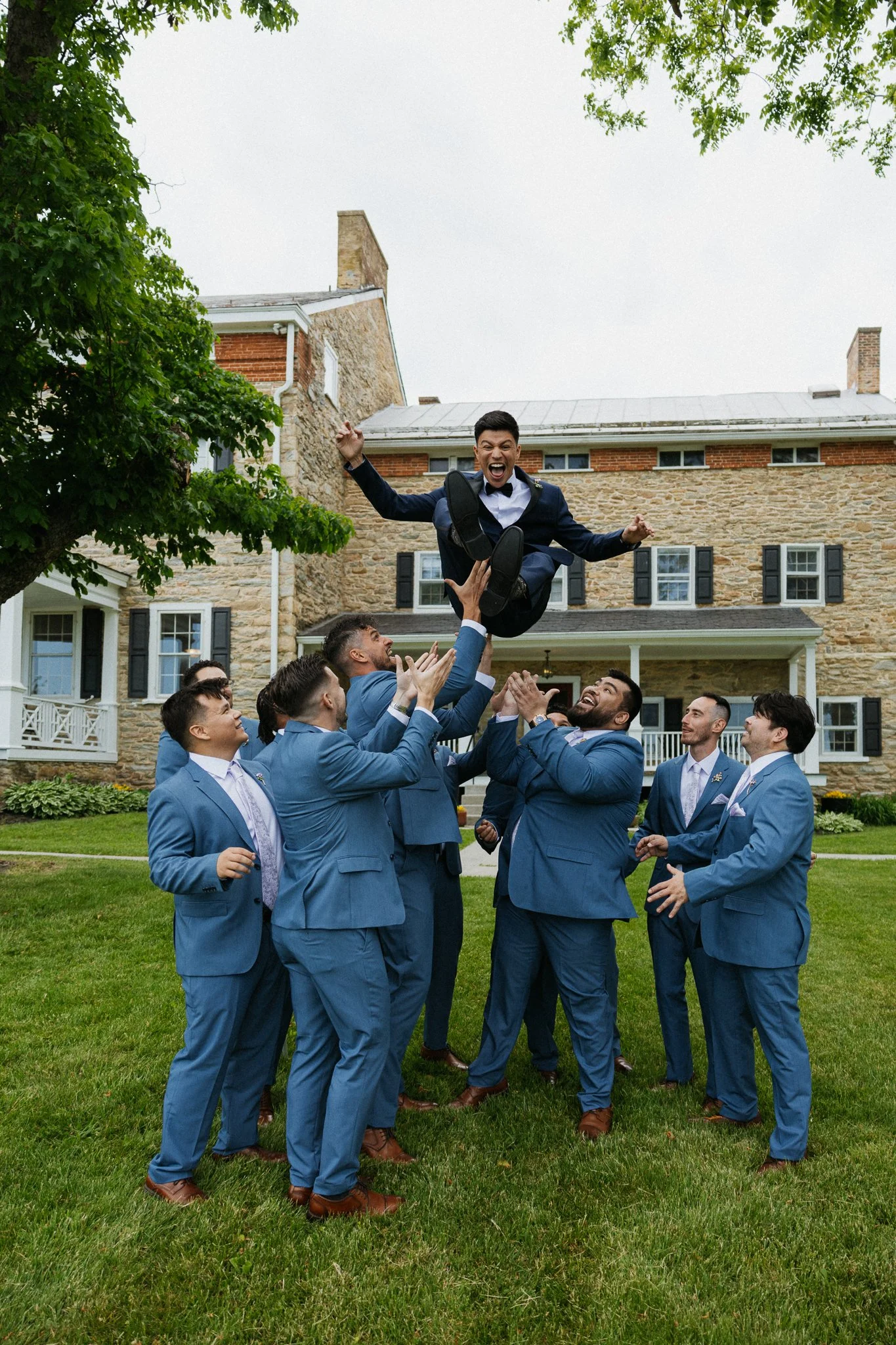 A group of men in blue suits having a outdoor celebration, with one man being lifted into the air, smiling and laughing, in front of a large house with a well-manicured lawn and trees.