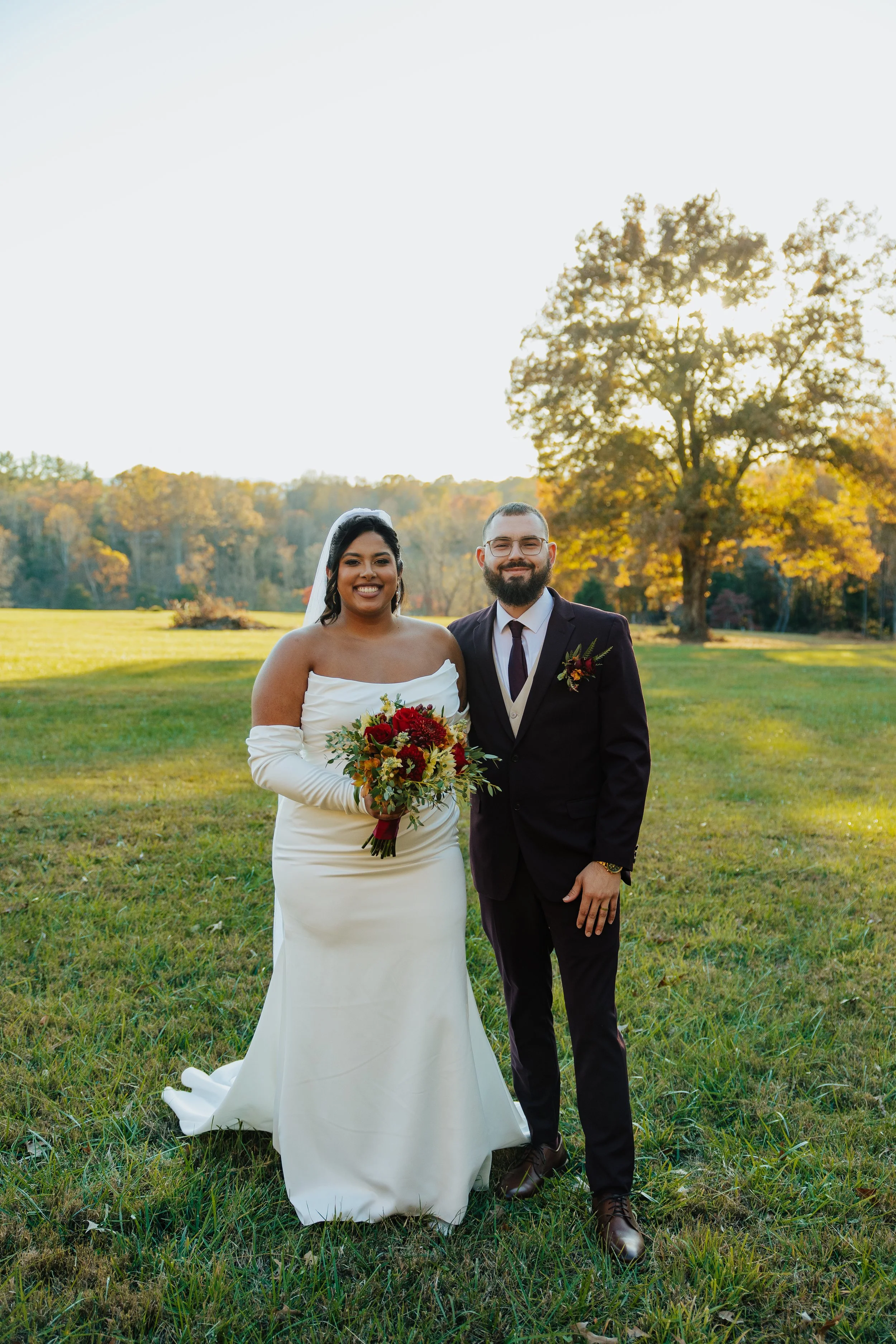 A bride and groom standing together outdoors, smiling, with trees in the background during a sunny day. The bride is holding a bouquet of flowers and is wearing a white wedding dress, while the groom is in a dark suit with glasses and a beard.