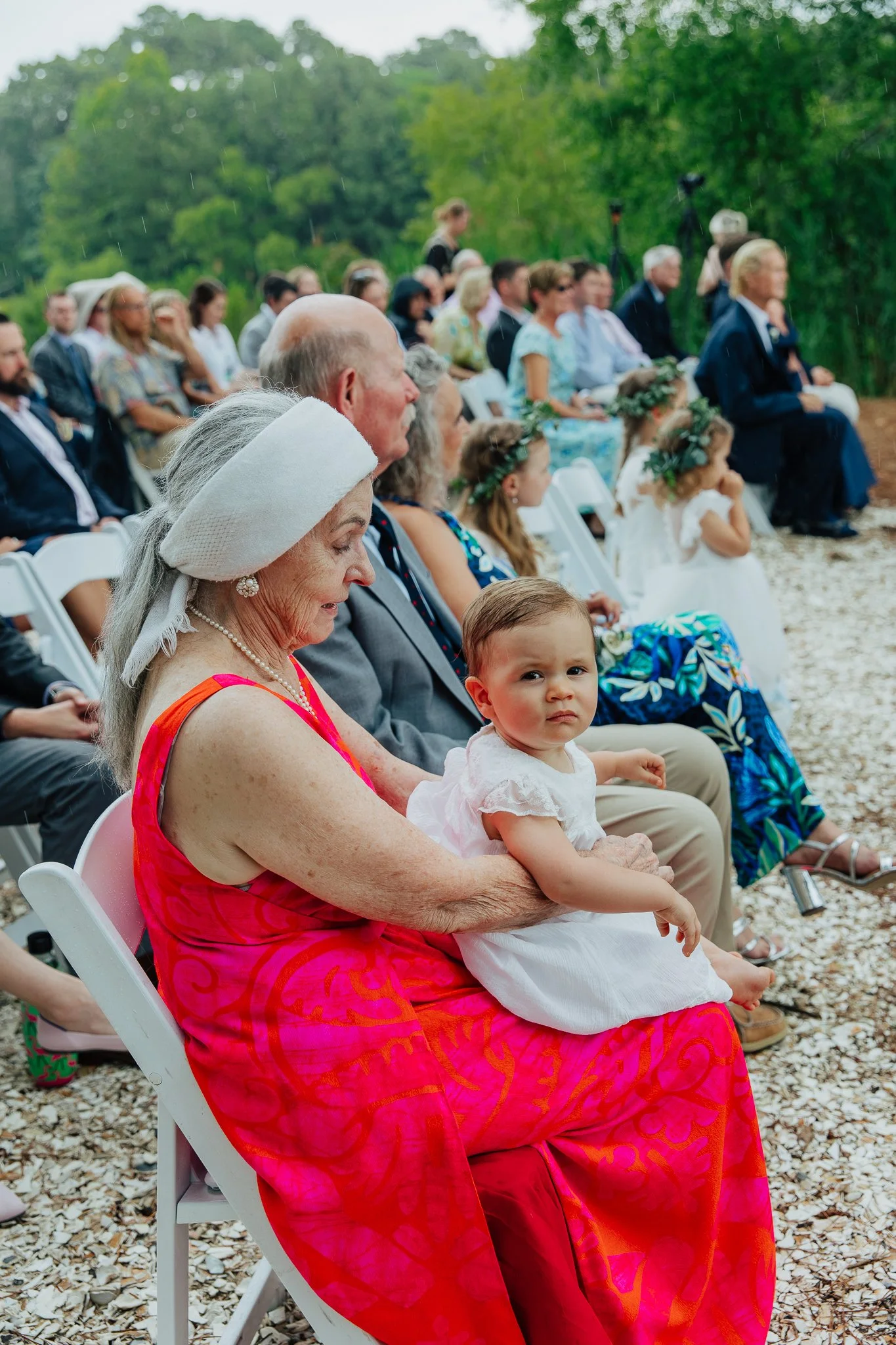 An elderly woman in a pink dress and white hat sitting outdoors on a white chair holding a young girl in a white dress, with a group of people seated behind them attending a wedding ceremony in the rain with greenery in the background.