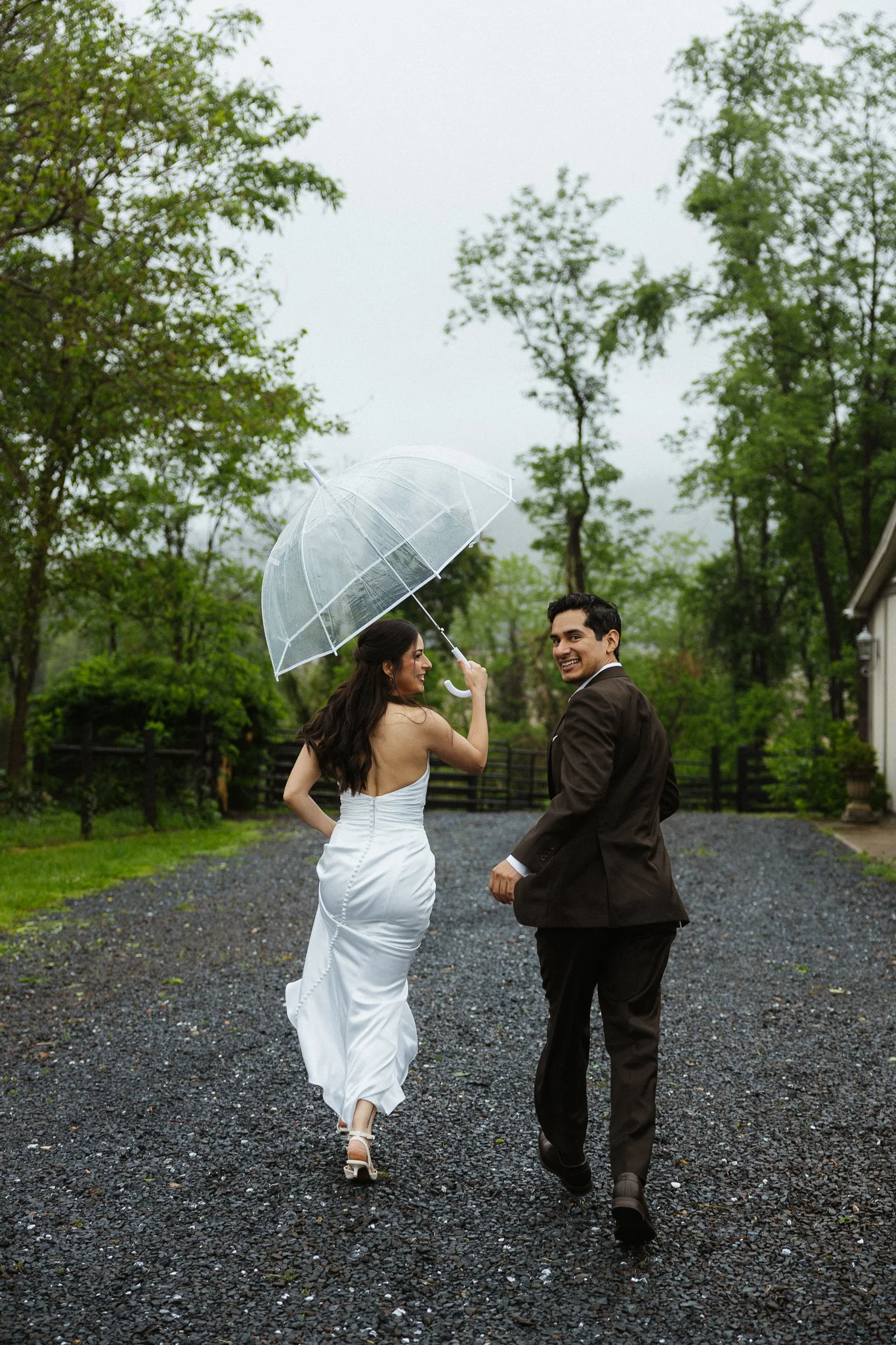 A bride and groom walking outdoors on a gravel path in the rain, holding a transparent umbrella, surrounded by green trees, smiling and looking happy.