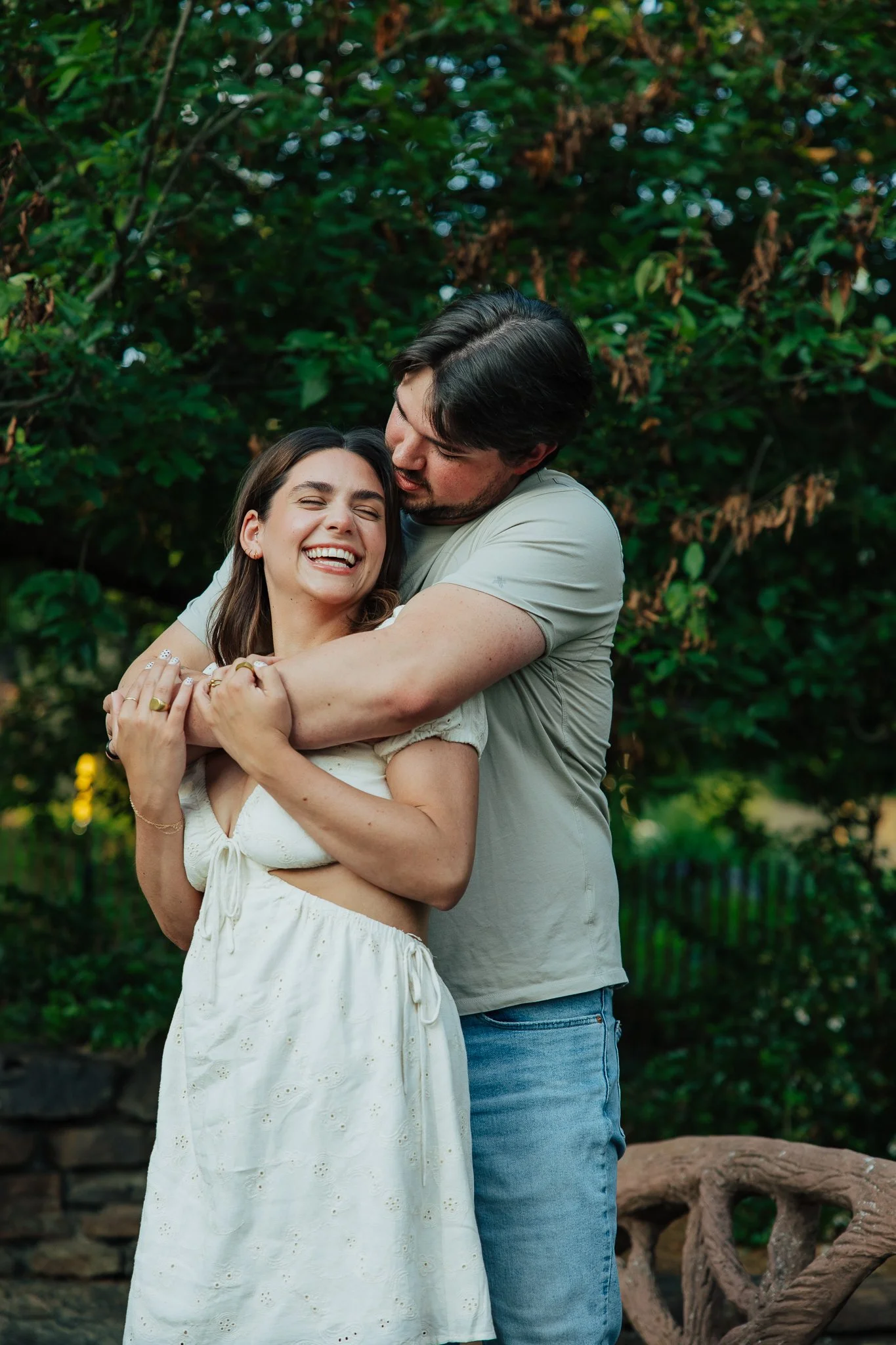 A couple happily embracing in an outdoor park or garden with green foliage in the background. The woman is smiling and the man is kissing her on the forehead.