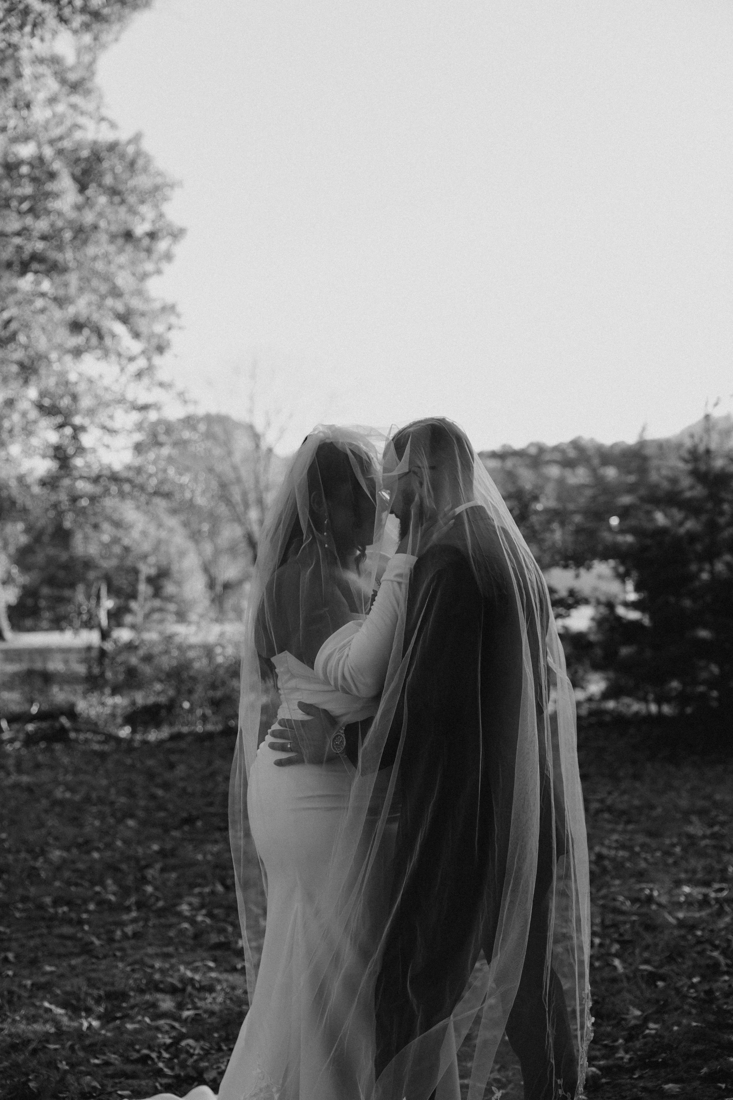 A black and white photo of two women in wedding dresses and veils standing close together outdoors, touching foreheads with their eyes closed.