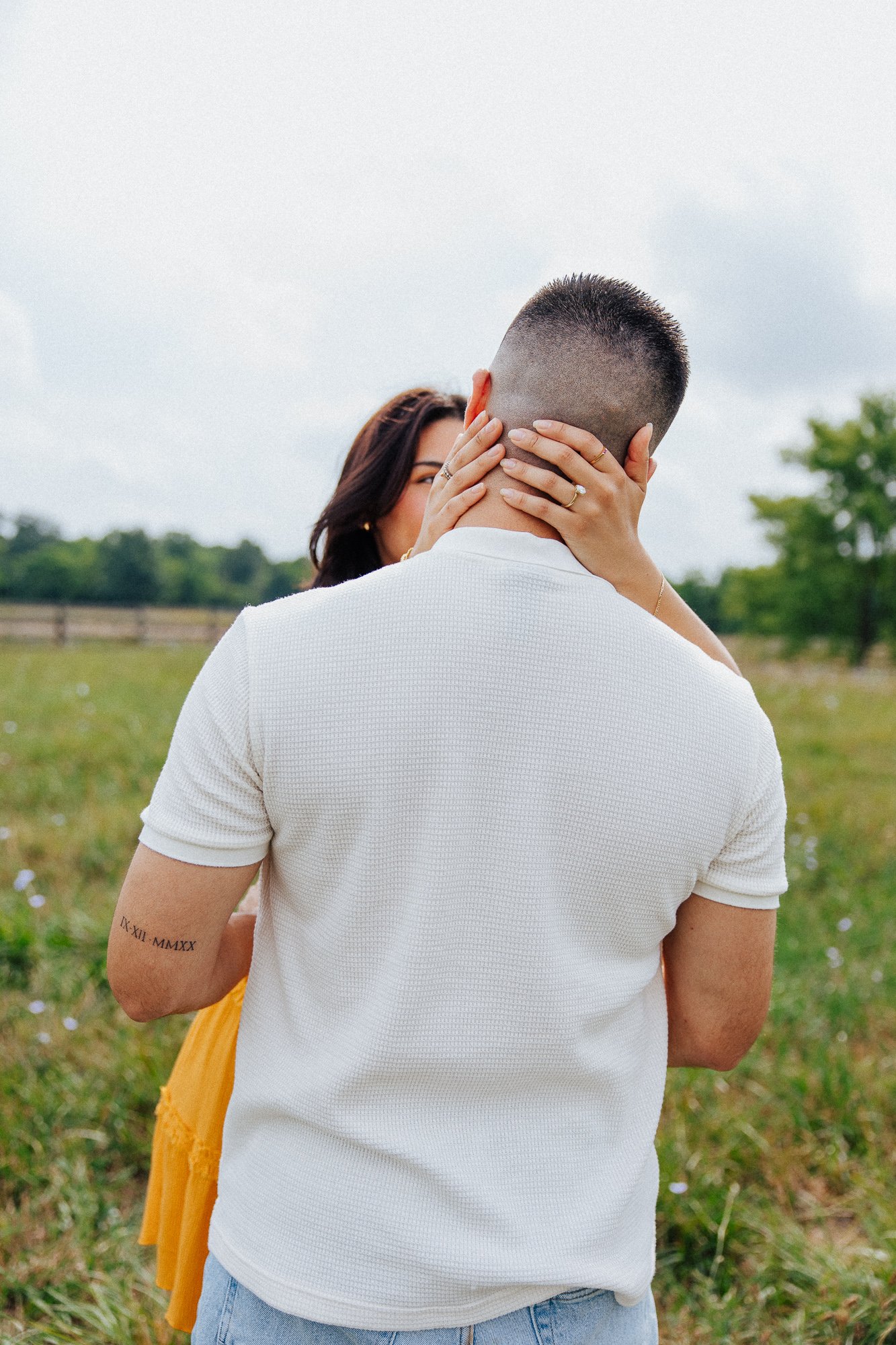 A man with a short haircut and tattoo on his arm stands outdoors. A woman with dark hair and jewelry holds his face with both hands, kissing him. The background shows an open field with trees and a cloudy sky.