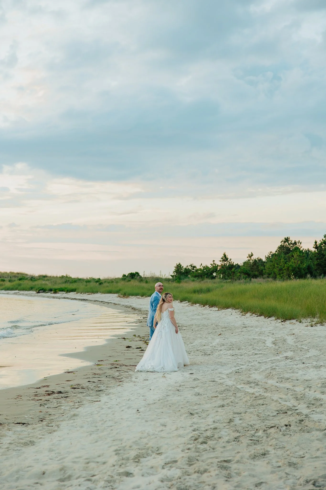 A bride and groom standing on the beach near the shoreline, with water and green trees in the background, during daytime.