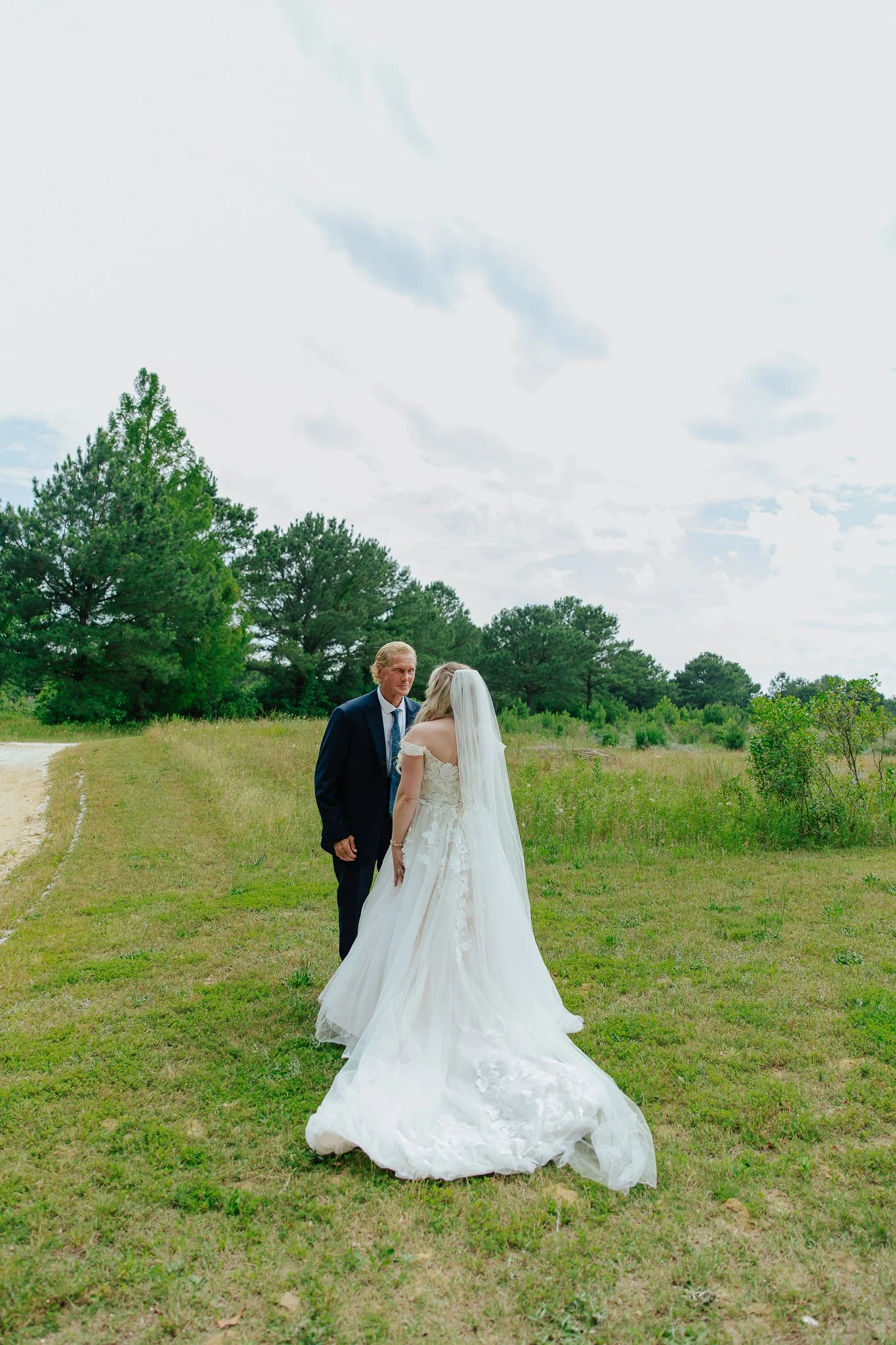 A bride and an older man, possibly her father, are standing on a grassy field with trees in the background during daytime. The groom is not visible. The bride is wearing a white wedding gown with lace details and a long veil.