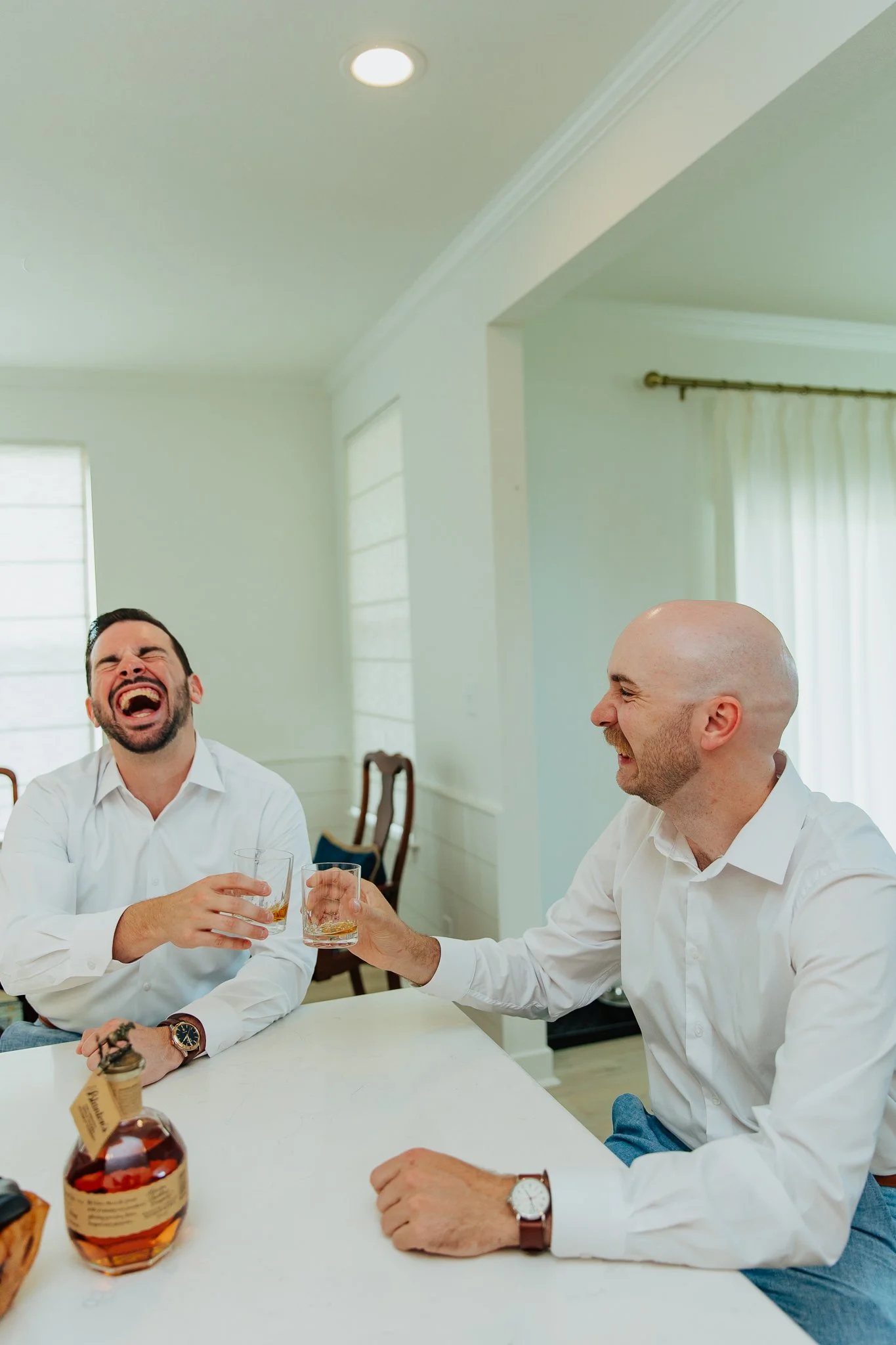 Two men in white shirts sitting at a table, sharing a toast with glasses of whiskey, smiling and laughing in a bright, modern room.