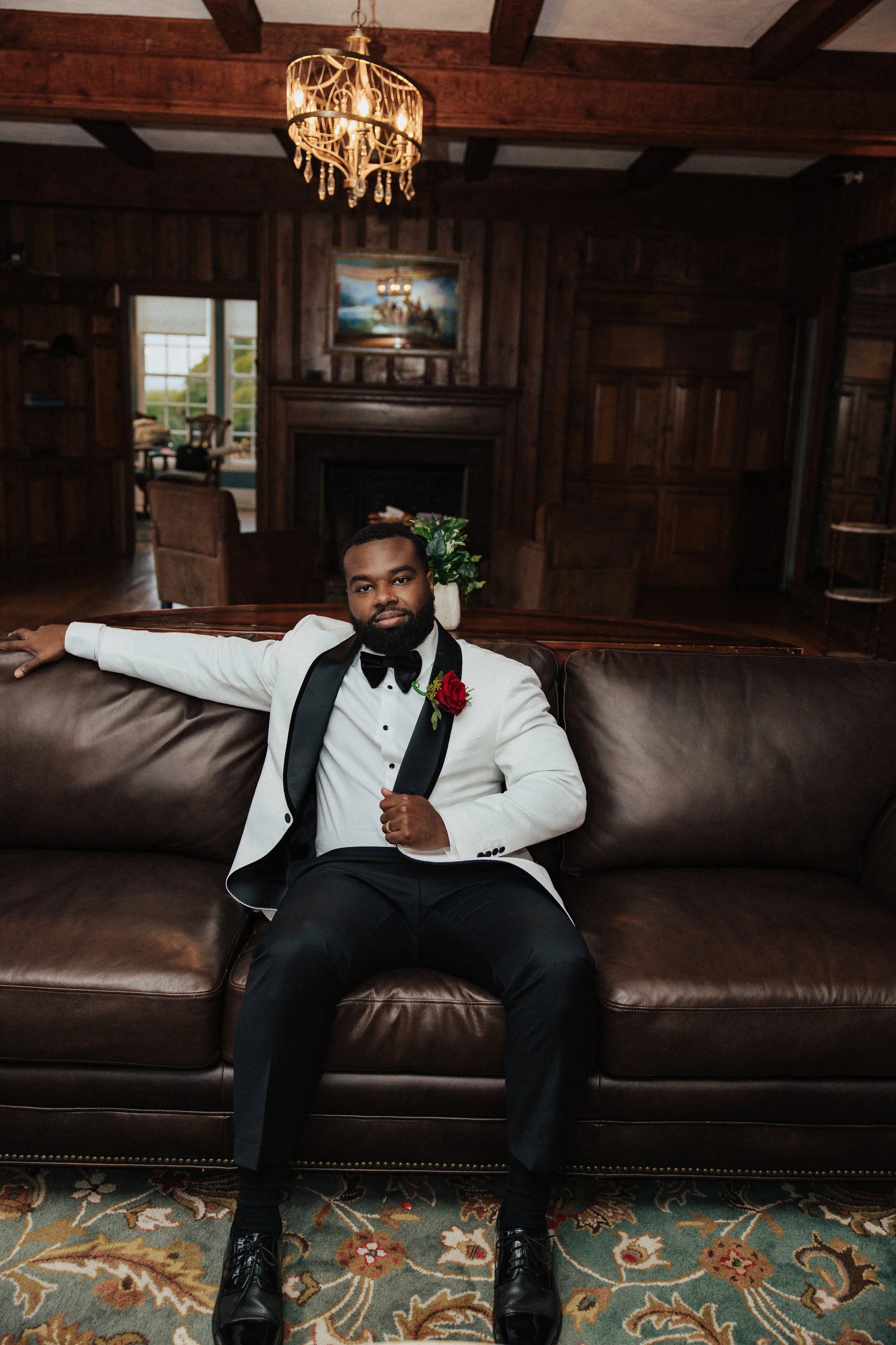 A man dressed in a white tuxedo jacket with black lapel, black pants, and black shoes, sitting on a brown leather sofa in a wood-paneled room, wearing a black bow tie and a red flower on his jacket, with his left arm resting on the back of the sofa a